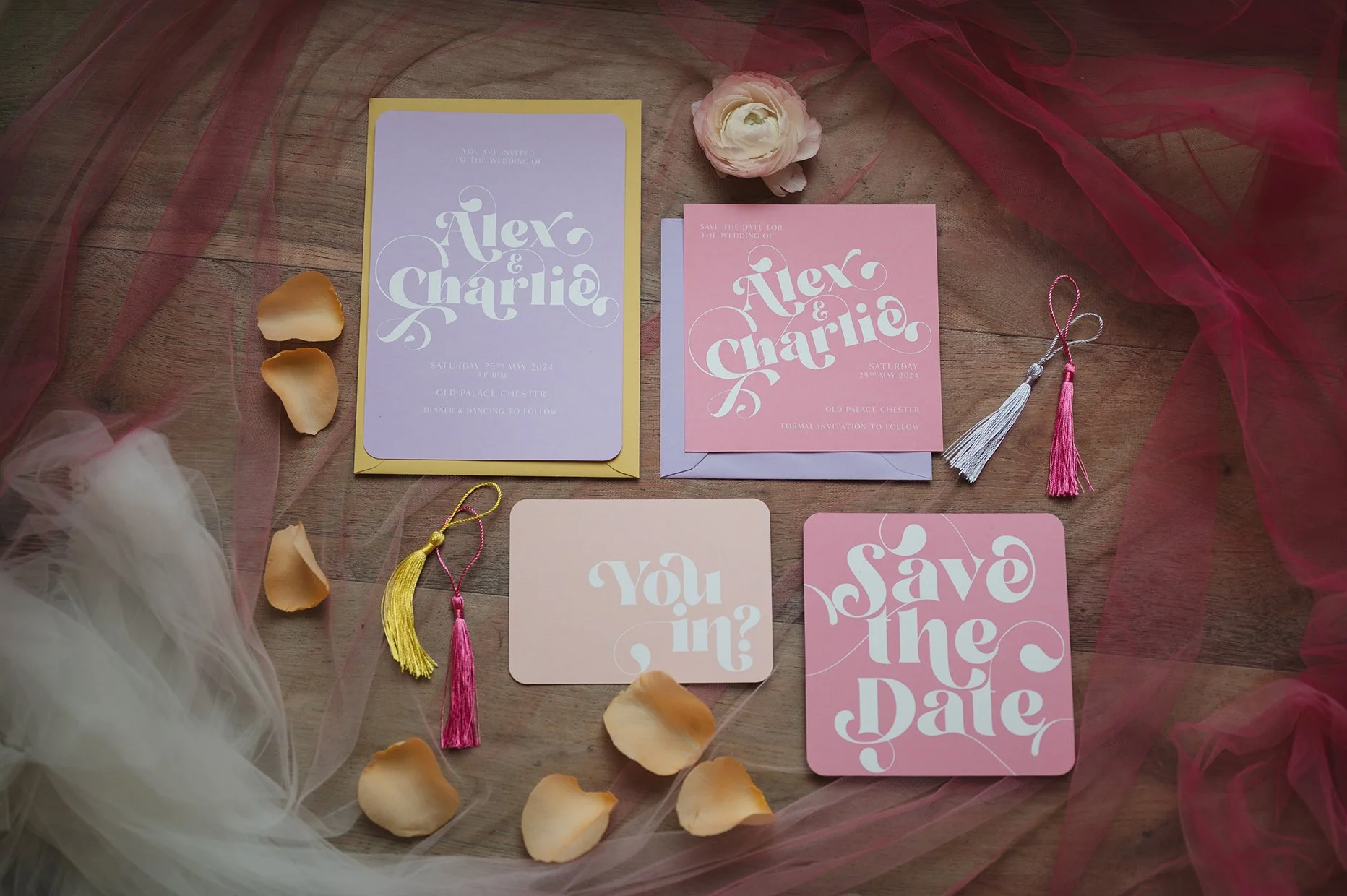 Wedding invitation cards for Alex and Charlie, including a purple, pink, and peach save the date card, a pink invitation card, and a peach RSVP card, all decorated with tassels, rose petals, and sheer fabric on a wooden surface.