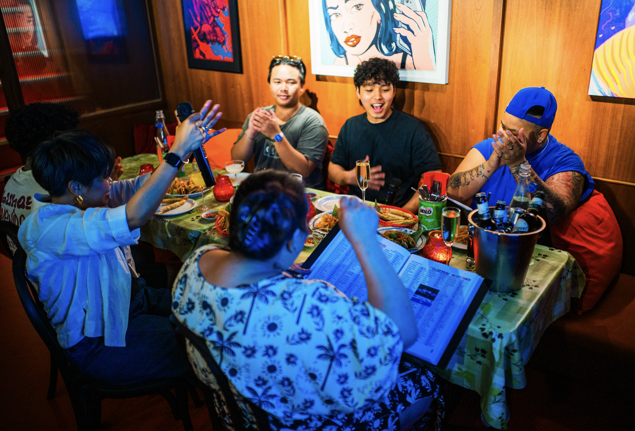 a table of people laughing and singing around a table of filipino food enjoying karaoke