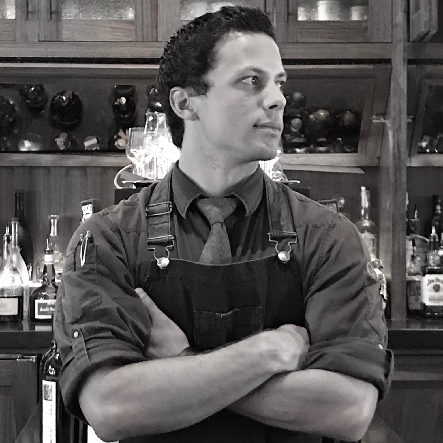 Black and white photo of Bartender Tanner Johnson standing in front of his bar with his arms crossed looking off camera.