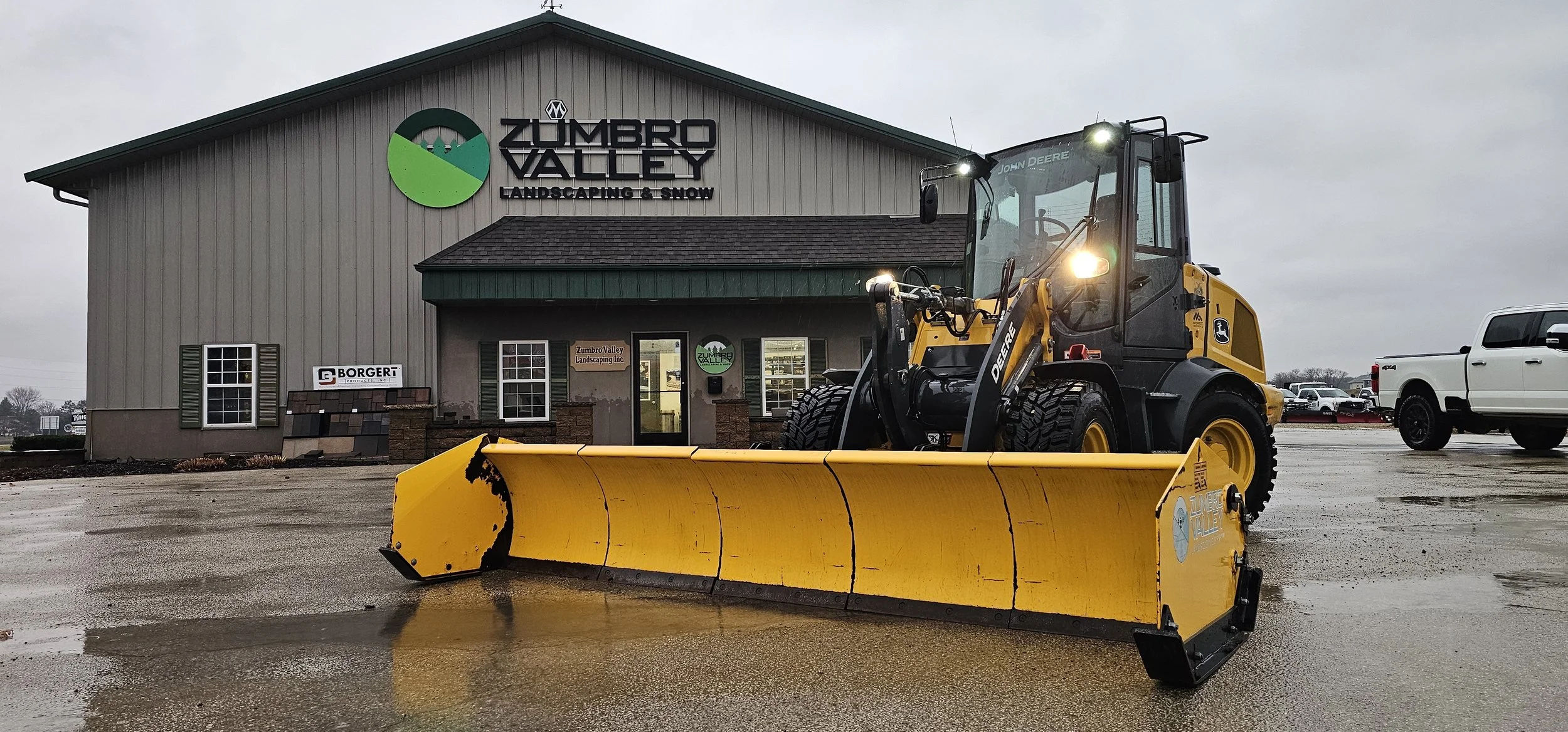 Snow-covered lot with two white trucks from Zumbro Valley Landscaping & Snow, one with a snowplow attachment, and a yellow skid steer loader, all parked in front of leafless trees and snow-covered ground.