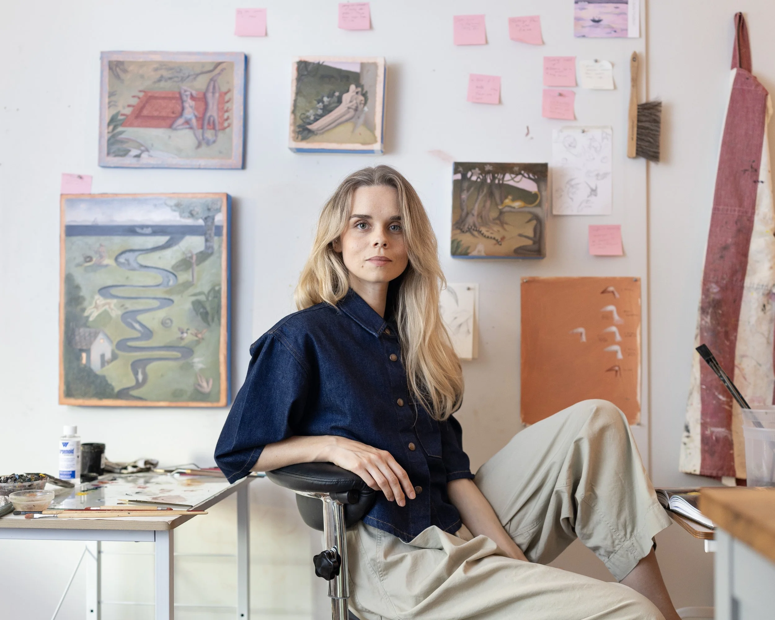 A young woman with blonde hair and a denim shirt sitting at an art studio desk with paintings and art supplies behind her, looking at the camera.