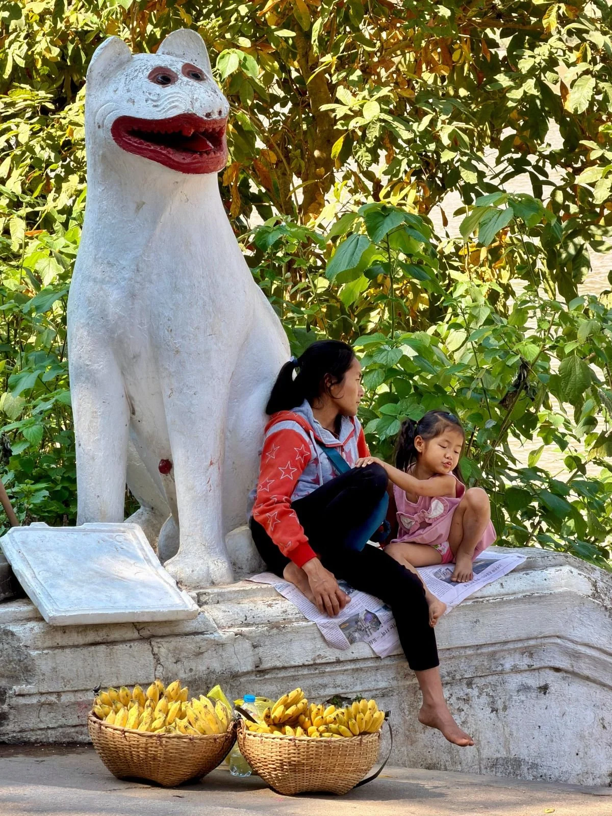 Laos - Rundreise auf eigene Faust - Bananenverkäuferin mit Kind unter einer Statue