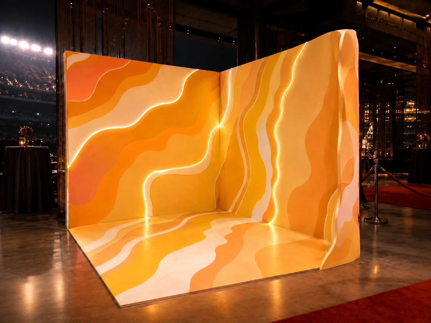 A wavy, orange and yellow, abstract wall with integrated lighting, creating a vibrant backdrop at an indoor event space, with black high-top tables and a red carpet separated by a velvet rope in the background.