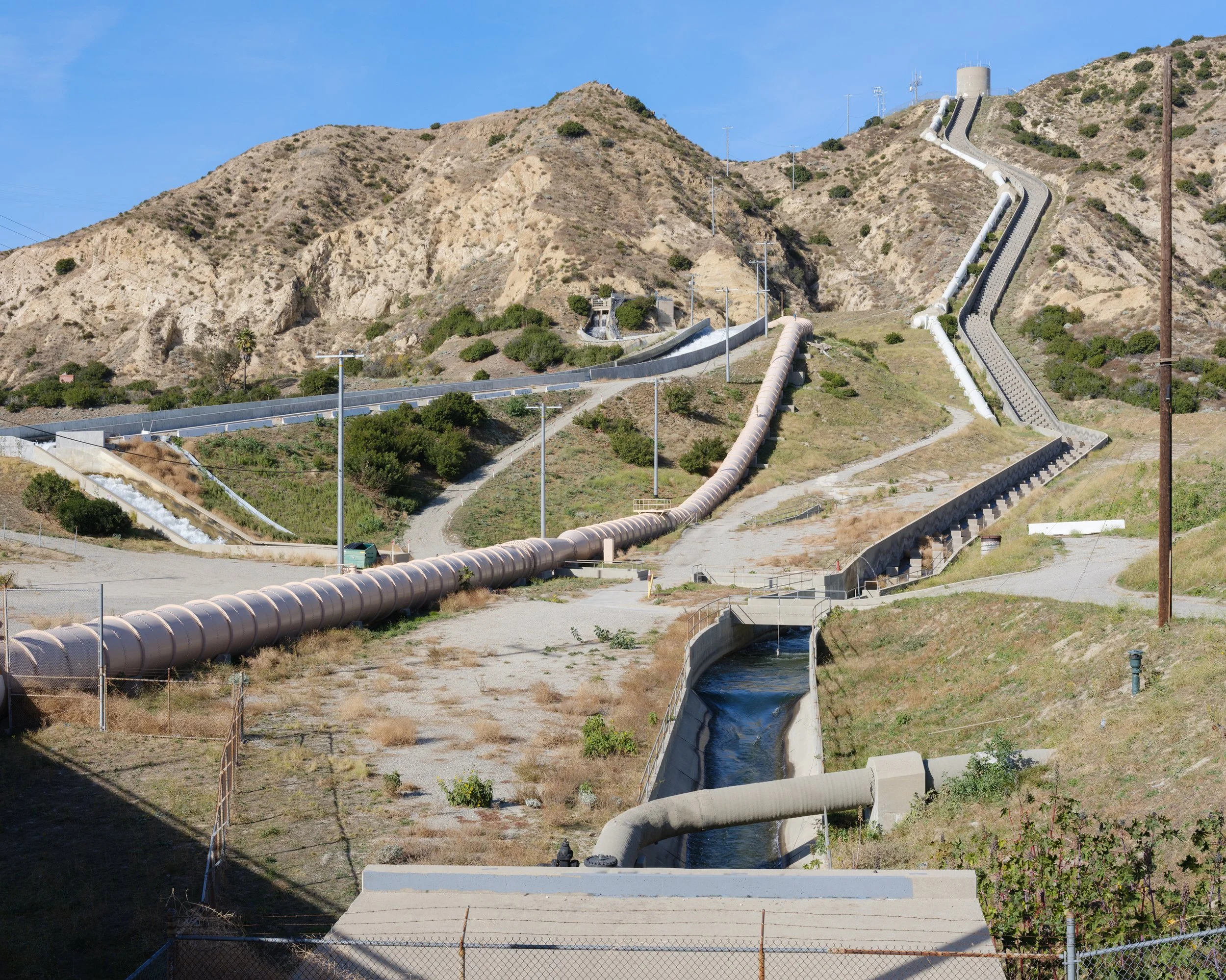 Water pipelines climb a dry hillside beneath a blue sky.