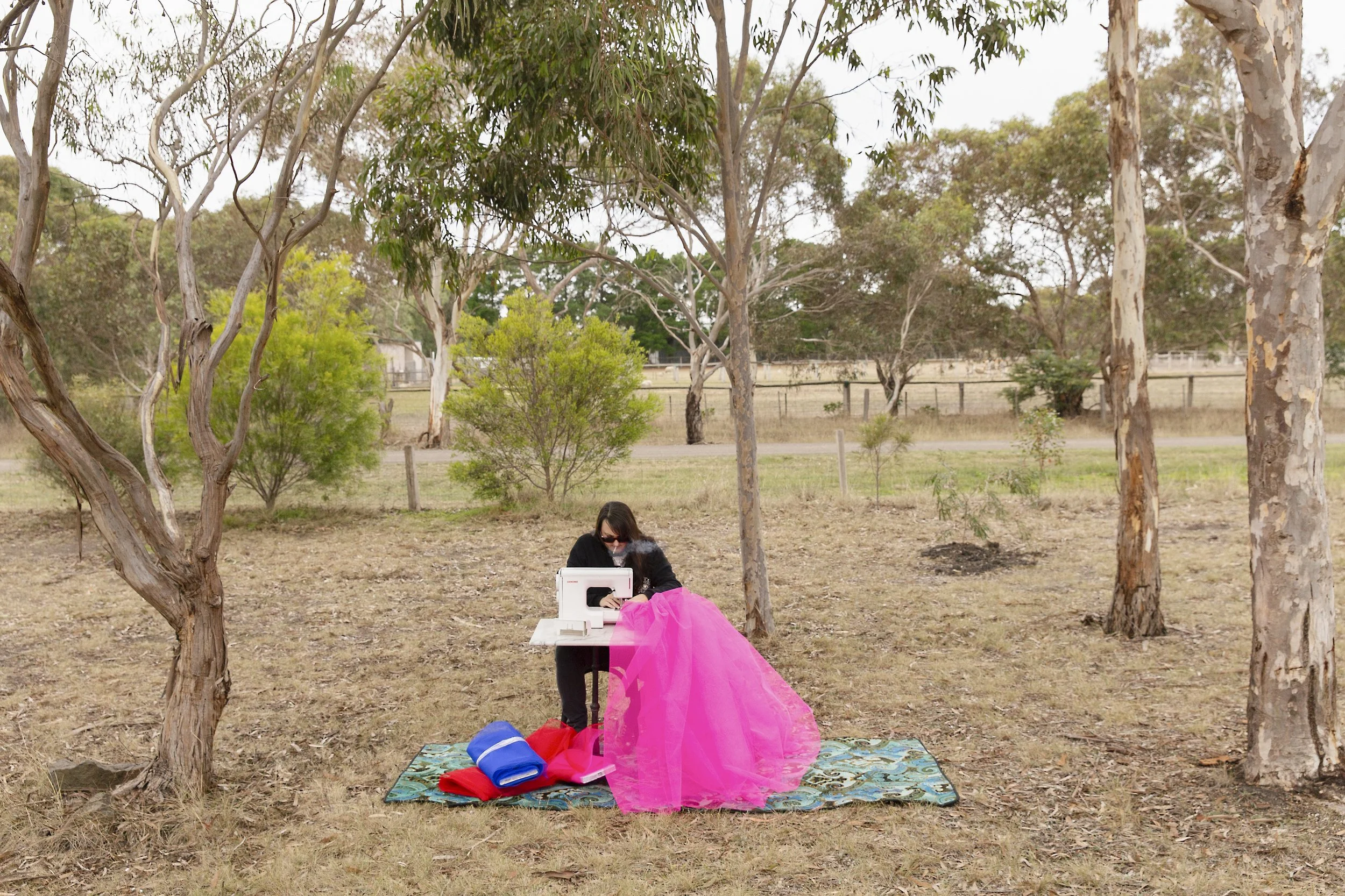 Artist sewing a pink fabric on a table outdoors in a park with trees and grass.