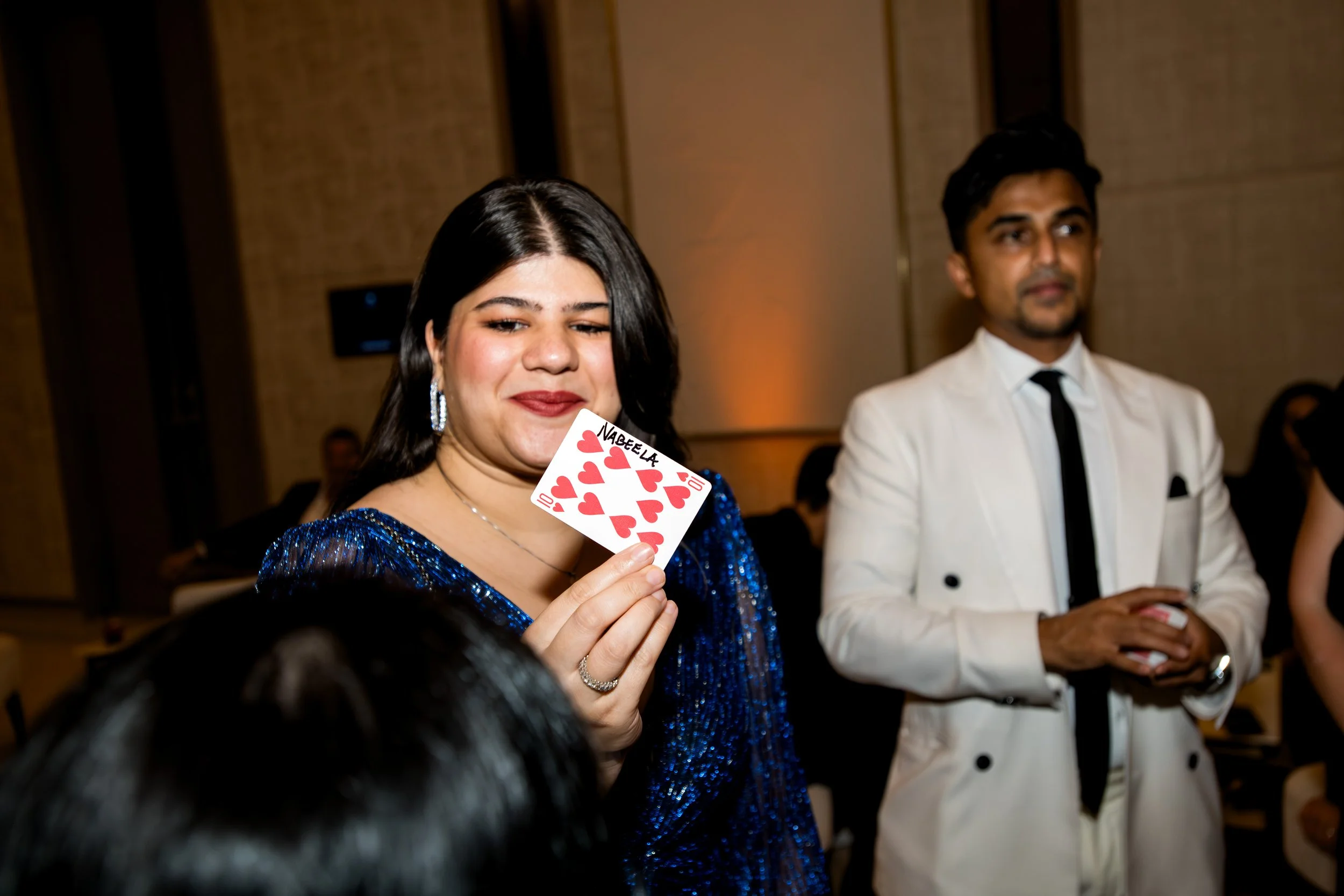 Magician in Dubai. Woman holding a playing card with seven hearts and the name 'Nabeela' written on it, at a formal event with a man in a white suit in the background.