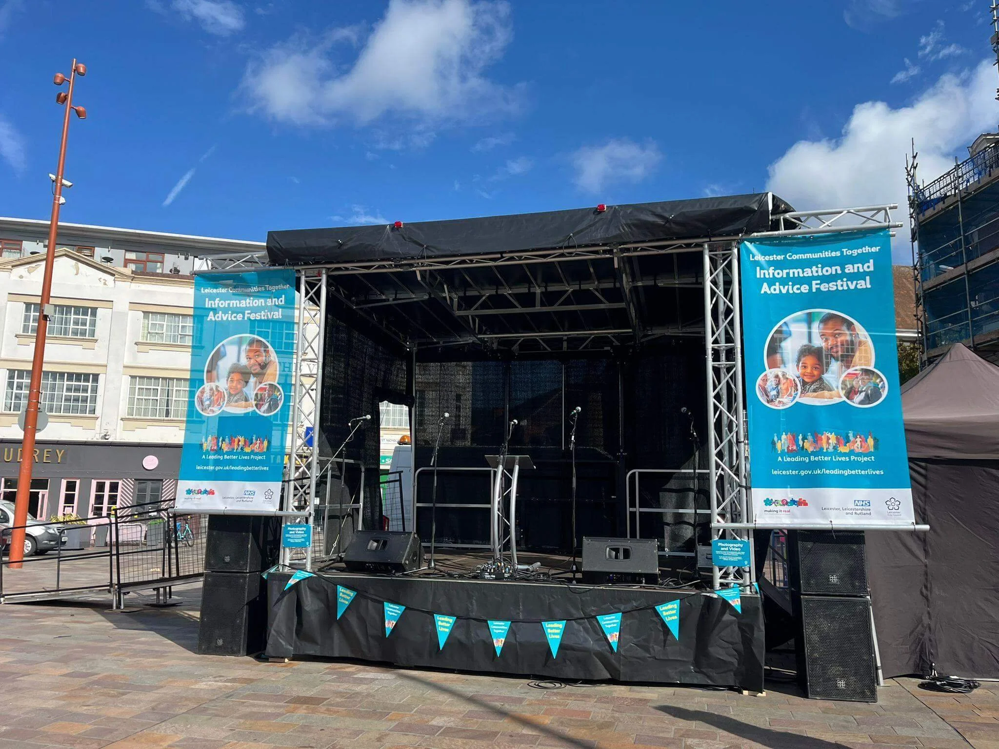 Outdoor mobile stage setup in city square with community festival banner branding, PA speakers, and lectern