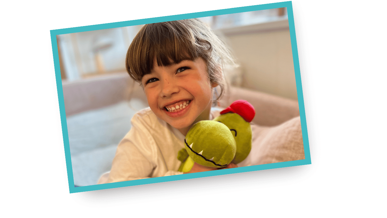 A young girl with brown hair holding a plush toy that looks like a green dinosaur with a red hat, smiling in a cozy indoor setting.