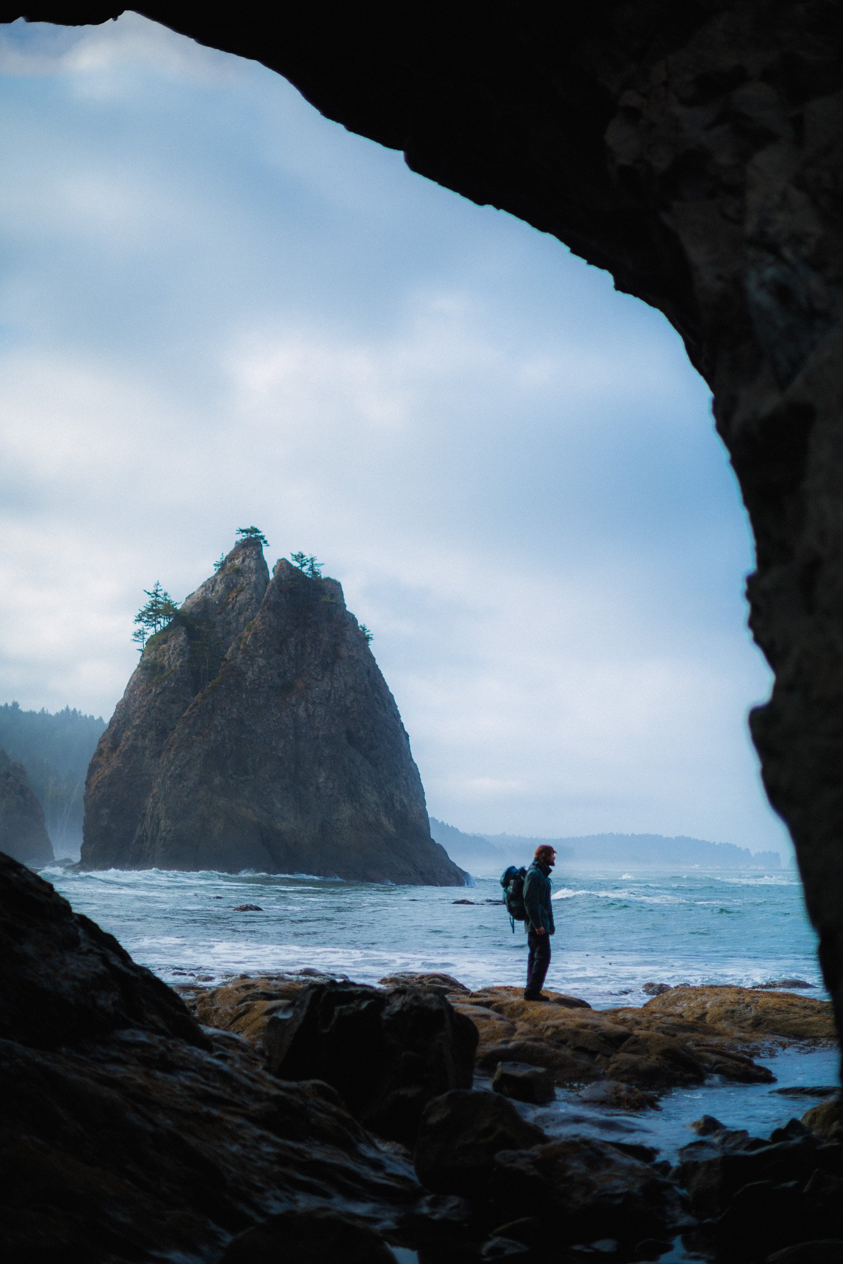 A person with a backpack standing on rocks near the ocean, looking at a large sea stack with trees on top, as viewed from inside a cave.