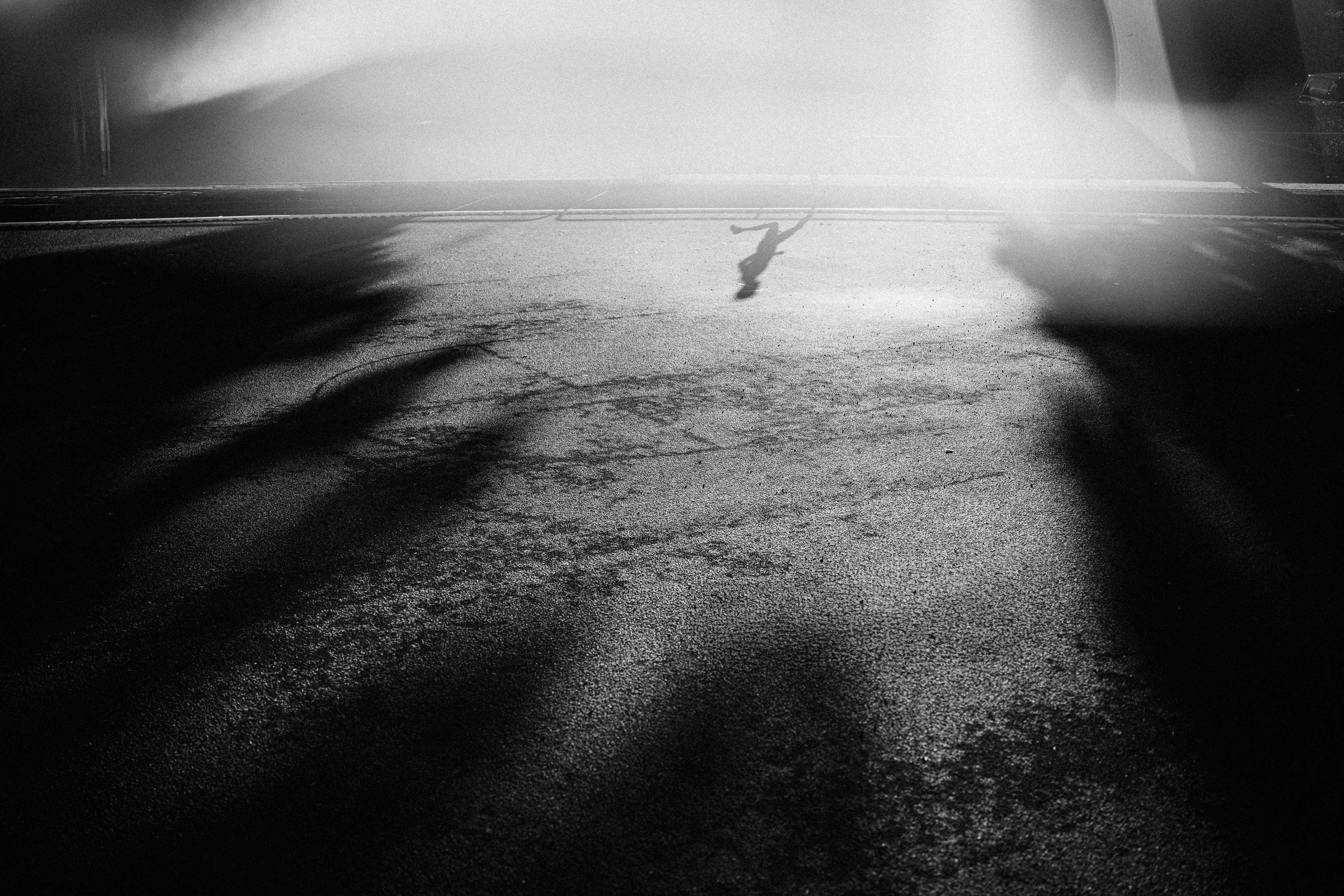 Silhouetted person walking on wet pavement with shadows and bright light glare, black and white photo