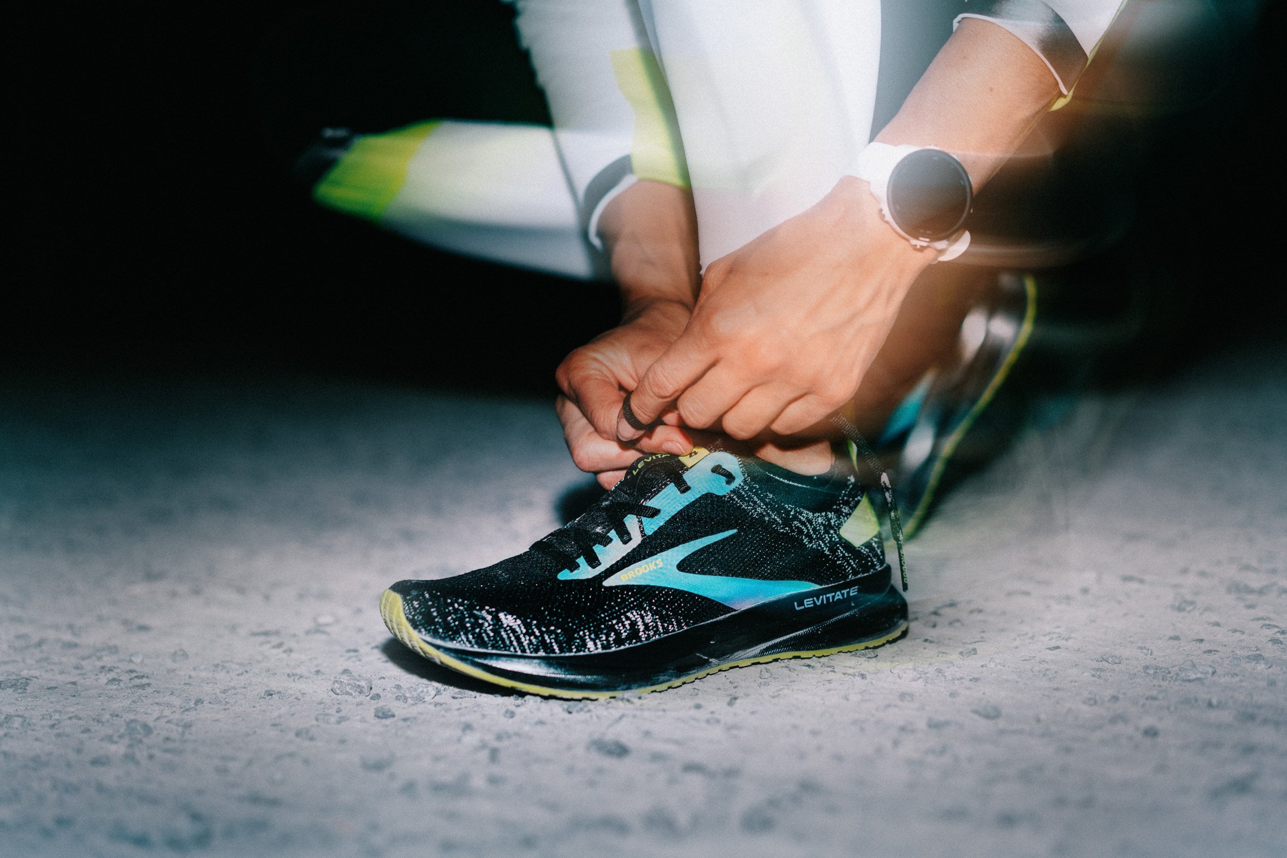 Person tying shoelace on a black running shoe with blue and yellow accents, on a gray surface.