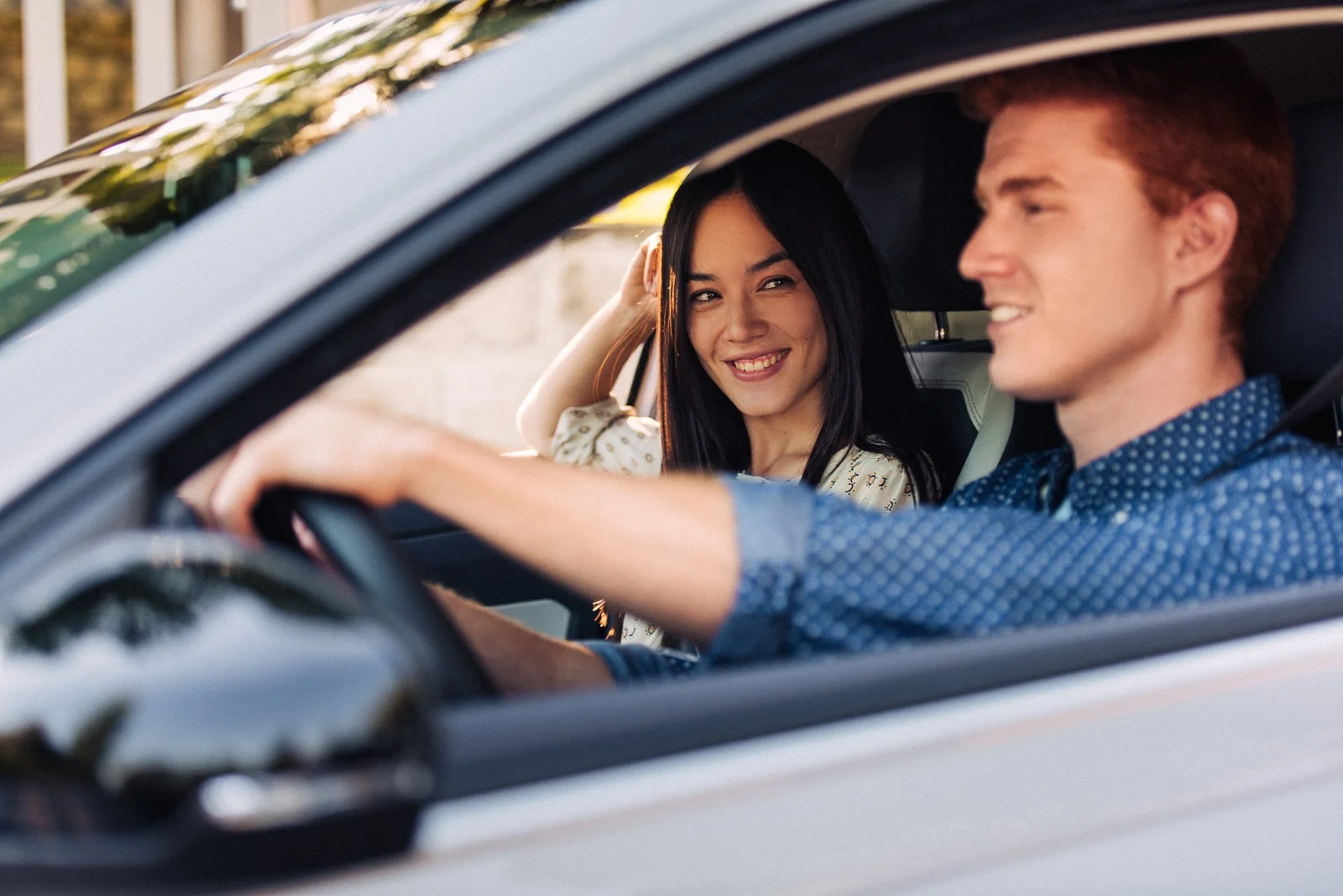 A young man and woman are sitting in a car; the man is driving and the woman is smiling and looking at the camera, with warm sunlight outside.
