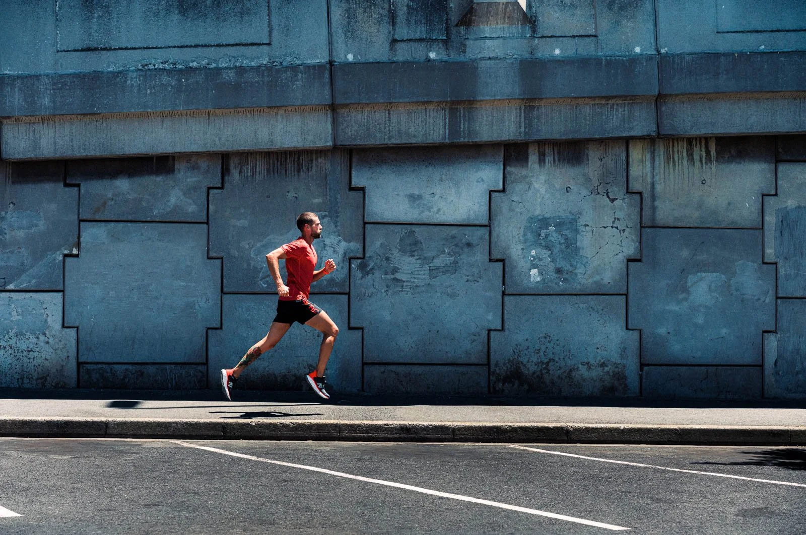 A man in a red shirt and black shorts running on a city street sidewalk with a blue textured wall in the background.