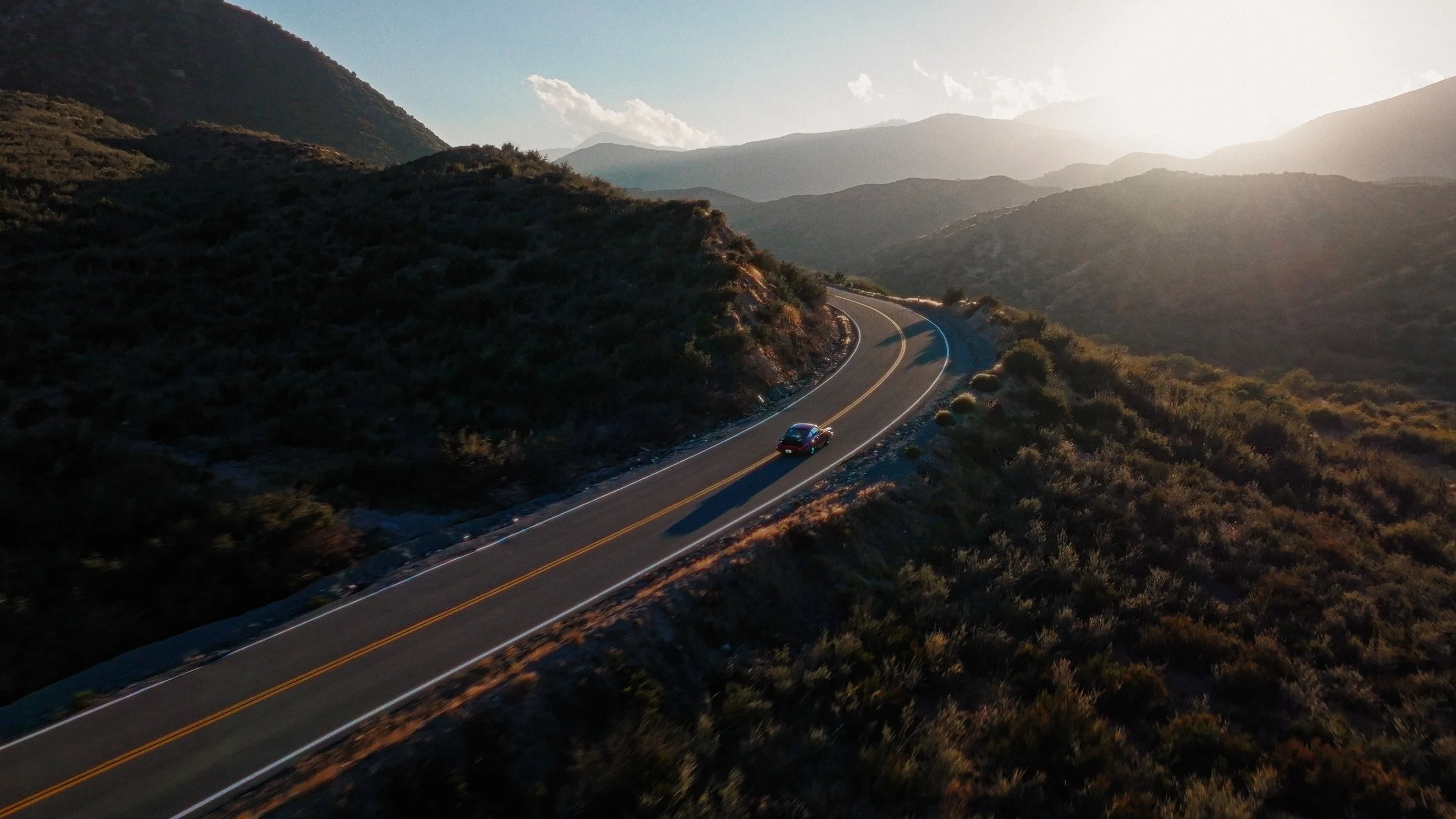 An aerial view of a winding mountain road with a single car driving in the late afternoon sunlight, surrounded by rolling hills and distant mountain ranges.