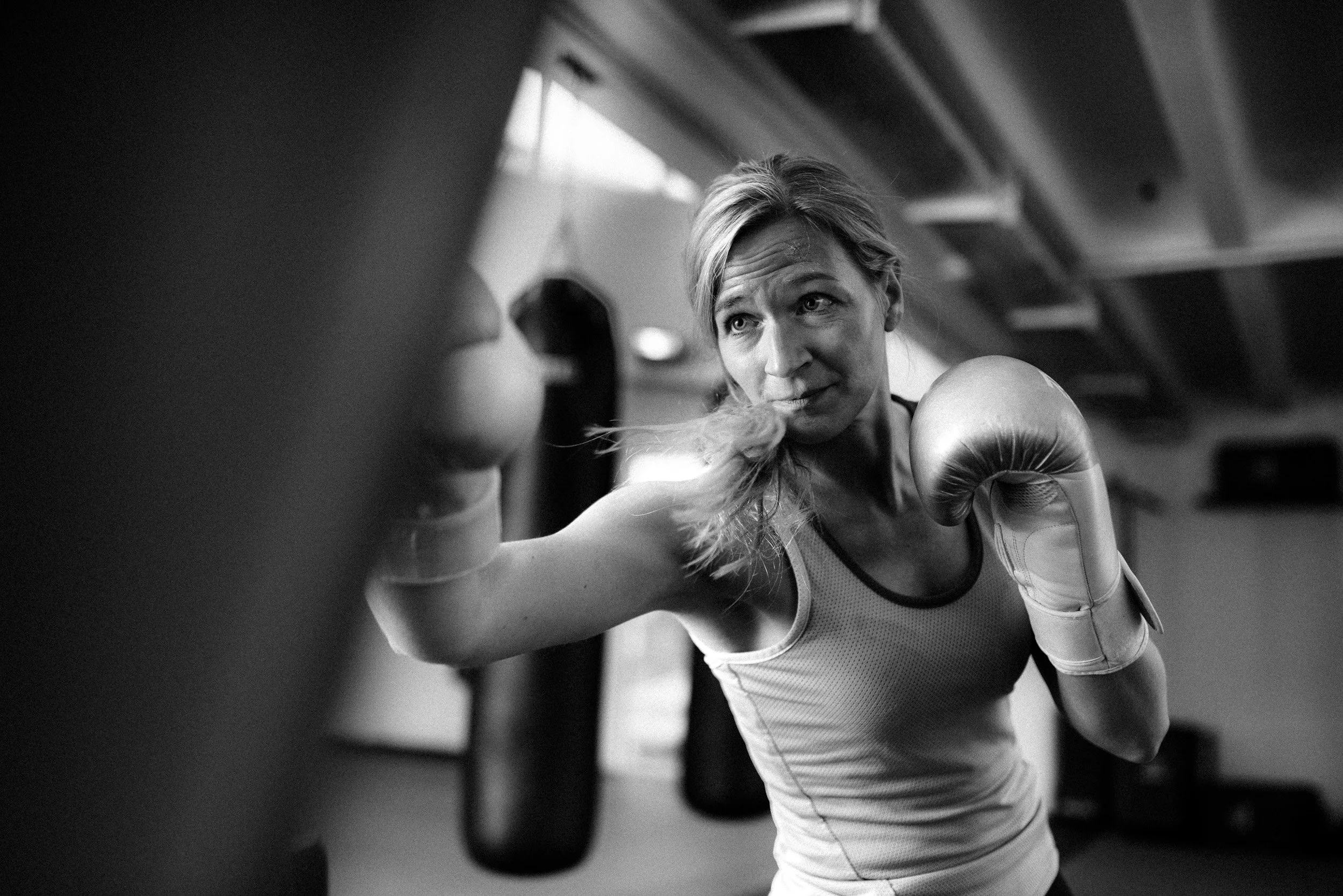 A woman wearing boxing gloves in a gym, leaning forward and punching a punching bag with a determined expression.