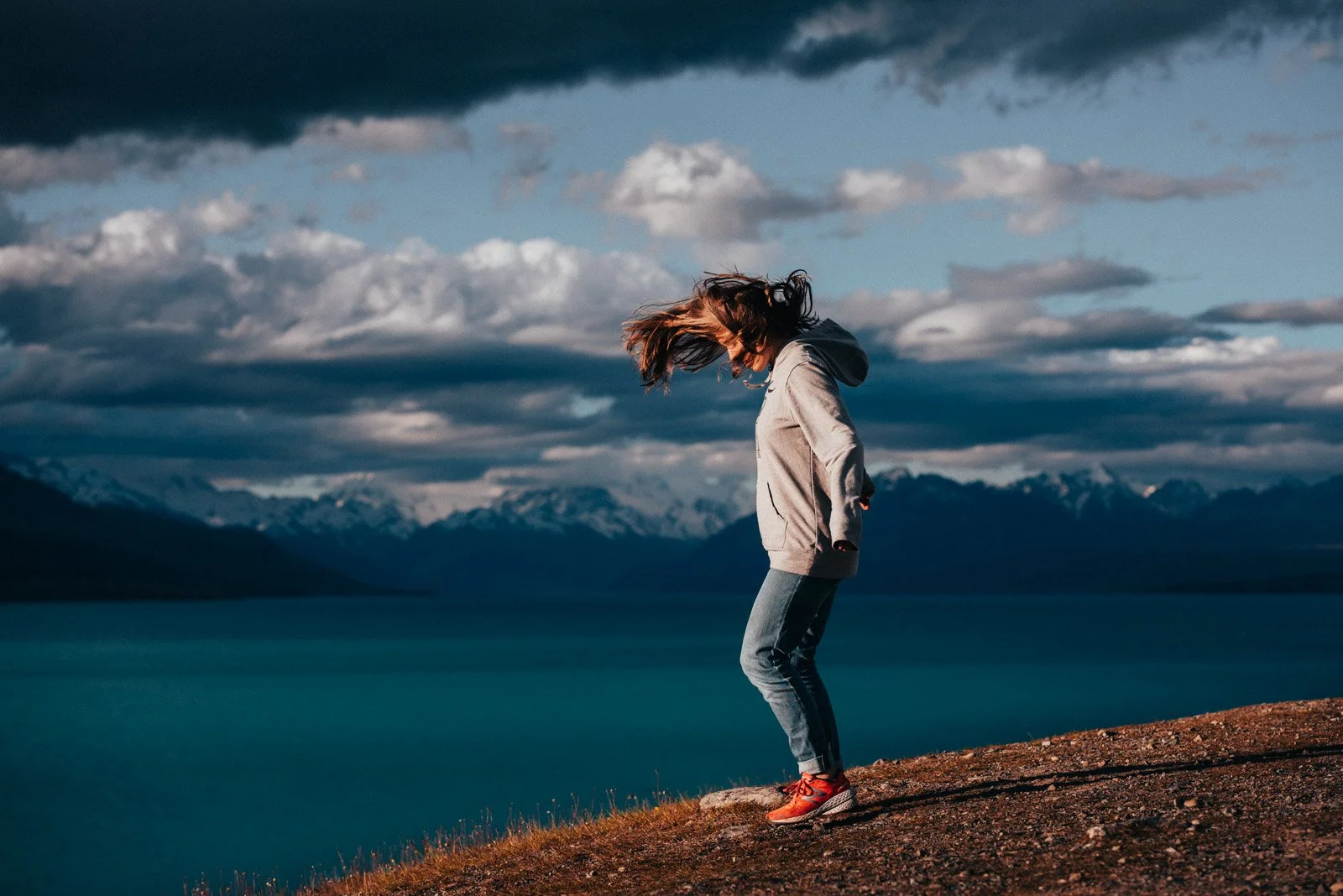 A woman with hair blowing in the wind is standing on a rocky hilltop near a body of water, with mountains in the distance beneath a cloudy sky.