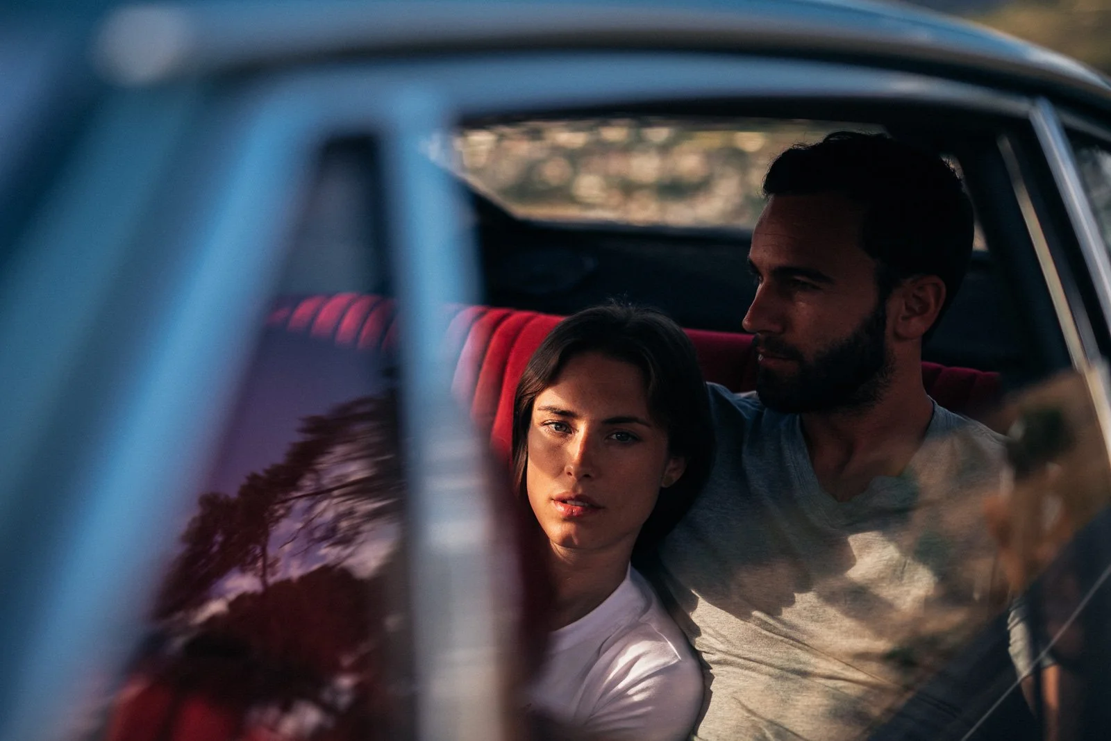 A young woman and a man sitting in the back seat of a car, seen through the window with reflections of trees and sky, during what appears to be late afternoon or early evening.