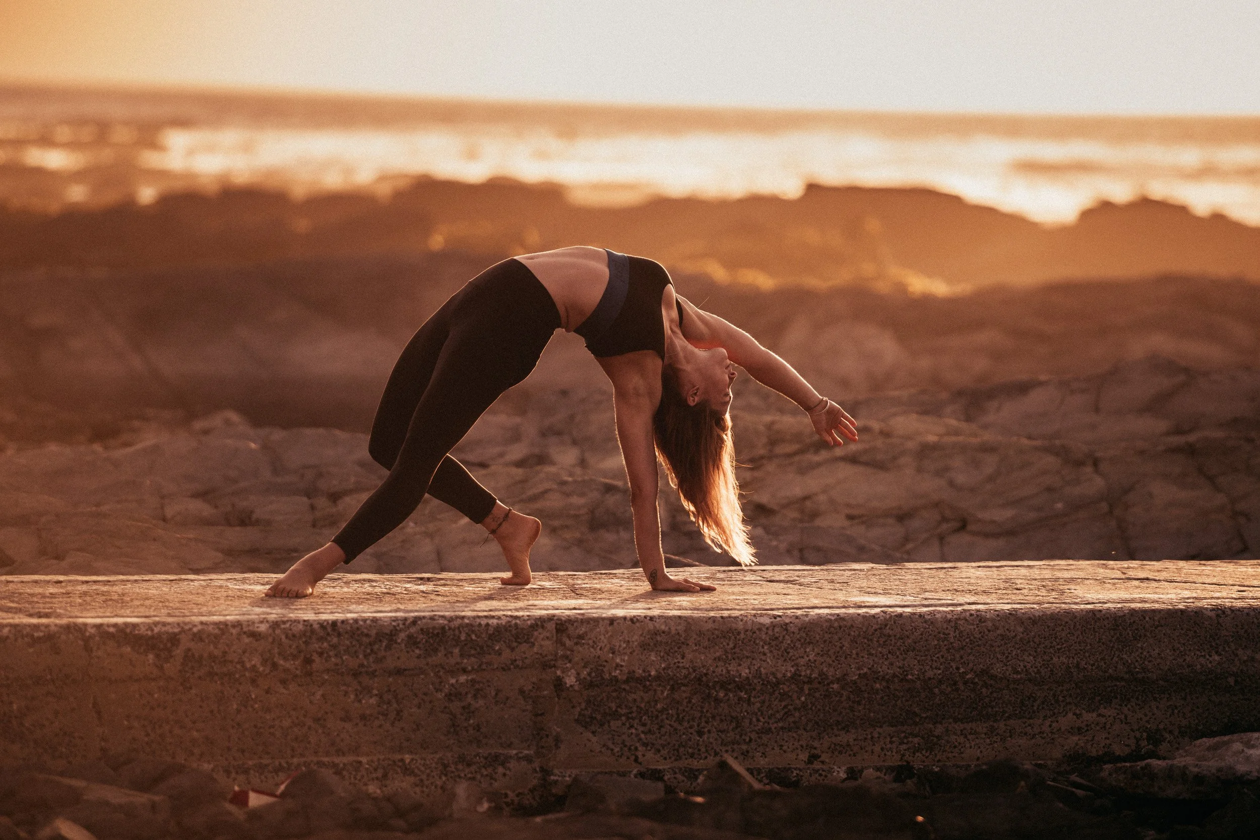Woman practicing yoga on a stone surface near the ocean at sunset.