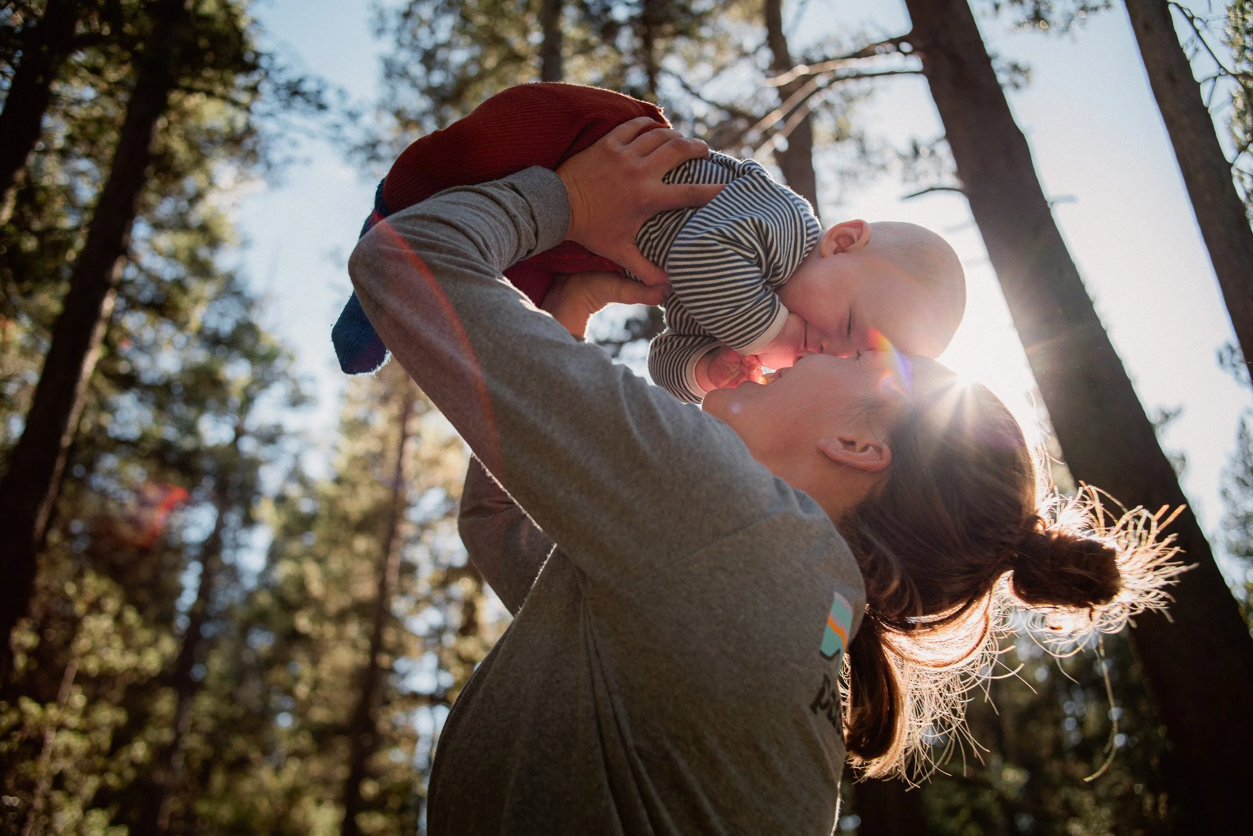 A woman lifting a baby outdoors in a forest with sunlight streaming through trees.