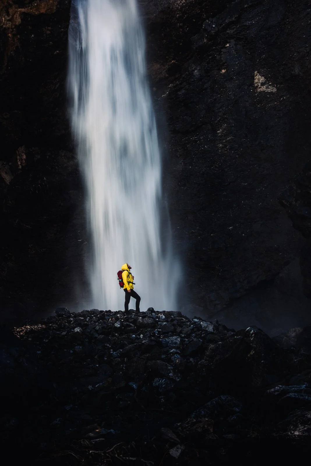 A person wearing a yellow jacket and hiking gear stands on dark rocks in front of a tall waterfall.