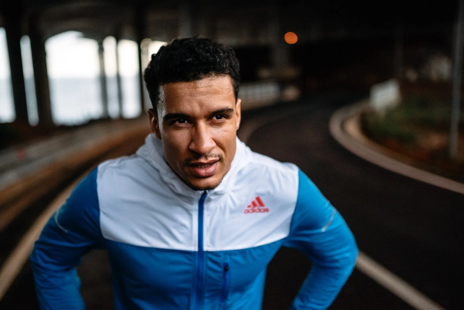 A man in a blue and white Adidas athletic jacket running outdoors under a bridge with curved railings