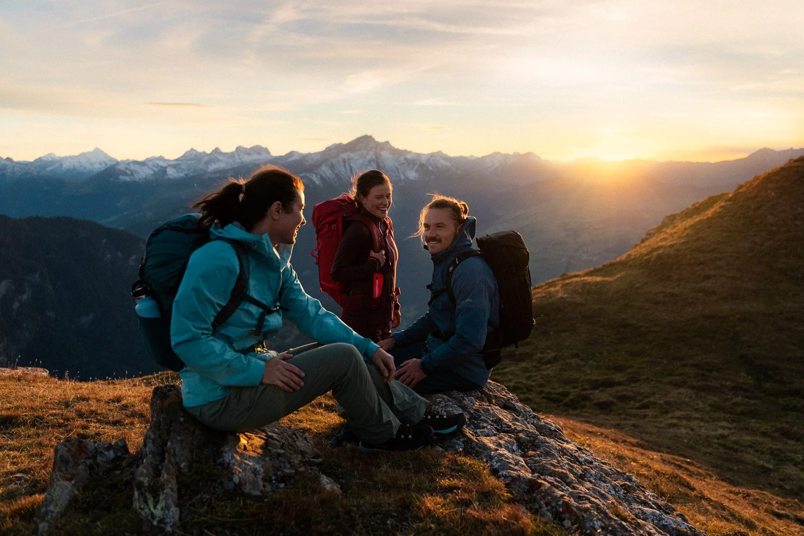 Four friends with backpacks enjoying a sunset view on a mountain top, sitting and standing on a rocky trail with mountains in the background.