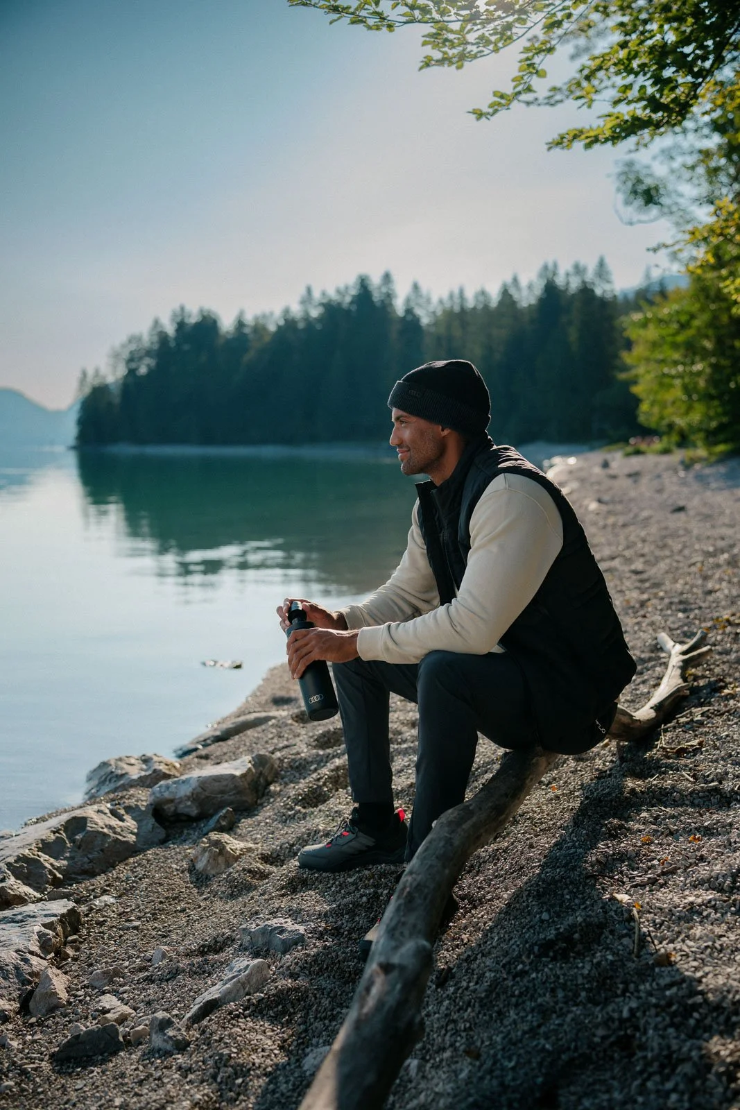 A man sitting on a log by a lake, holding a water bottle, wearing outdoor clothes and a beanie, with trees and mountains in the background.