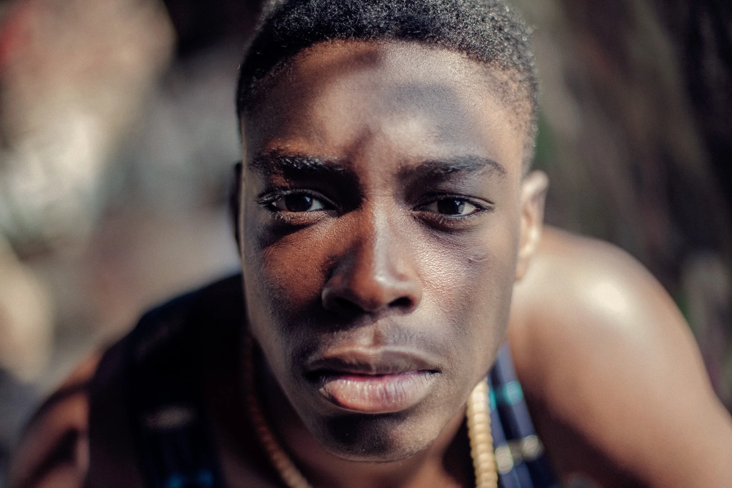 Close-up of a young man with short hair, wearing traditional attire and beads, looking into the camera with a serious expression