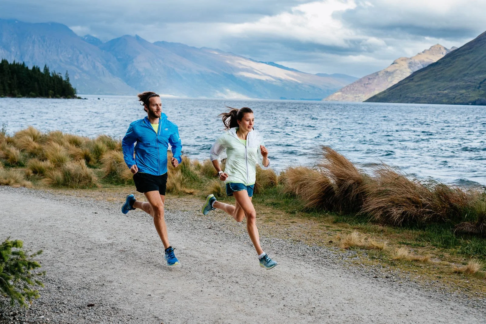 A man and woman running along a lakeshore with mountains in the background, wearing athletic clothing.