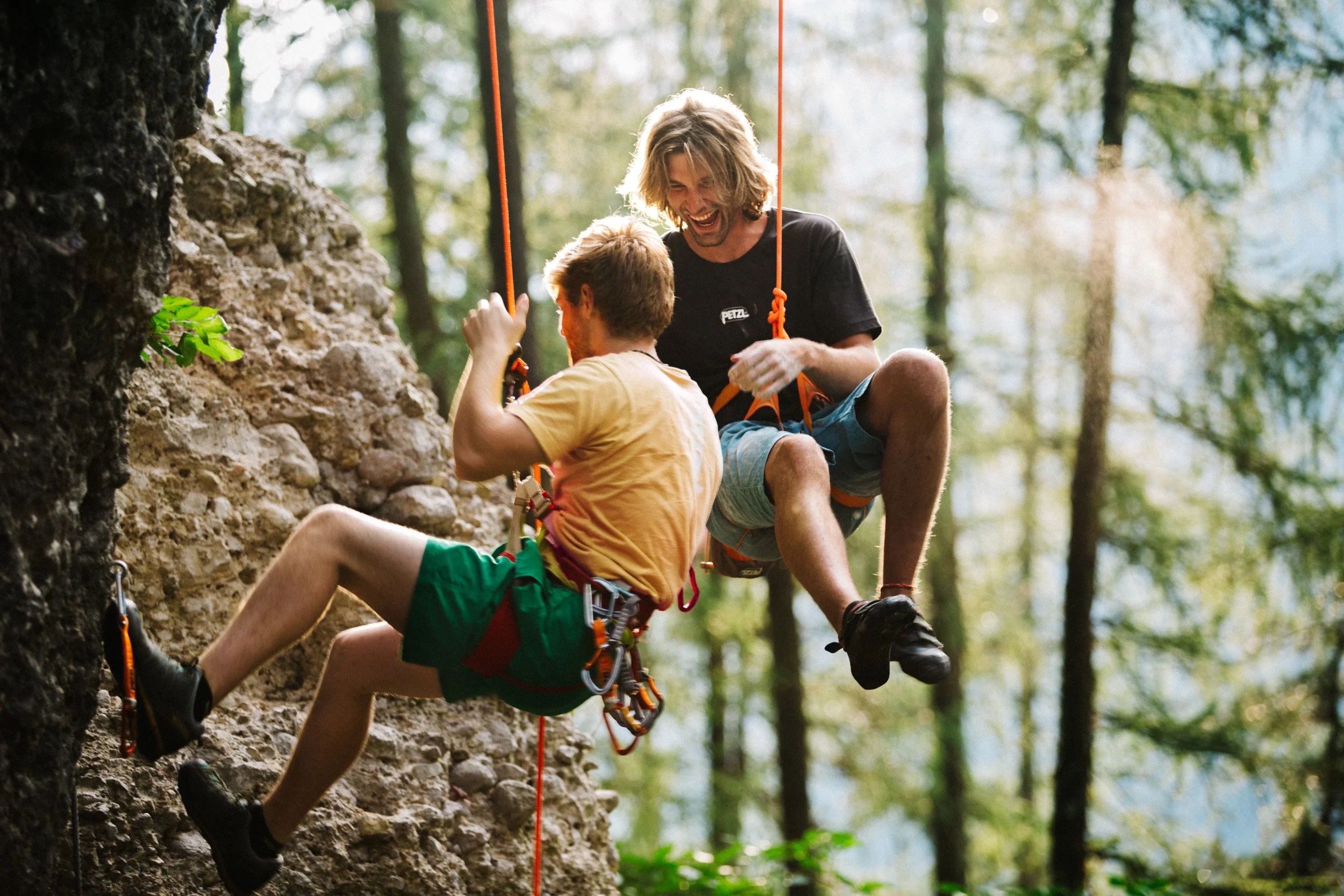 Two people rock climbing and laughing on a cliff outdoors, one helping the other.