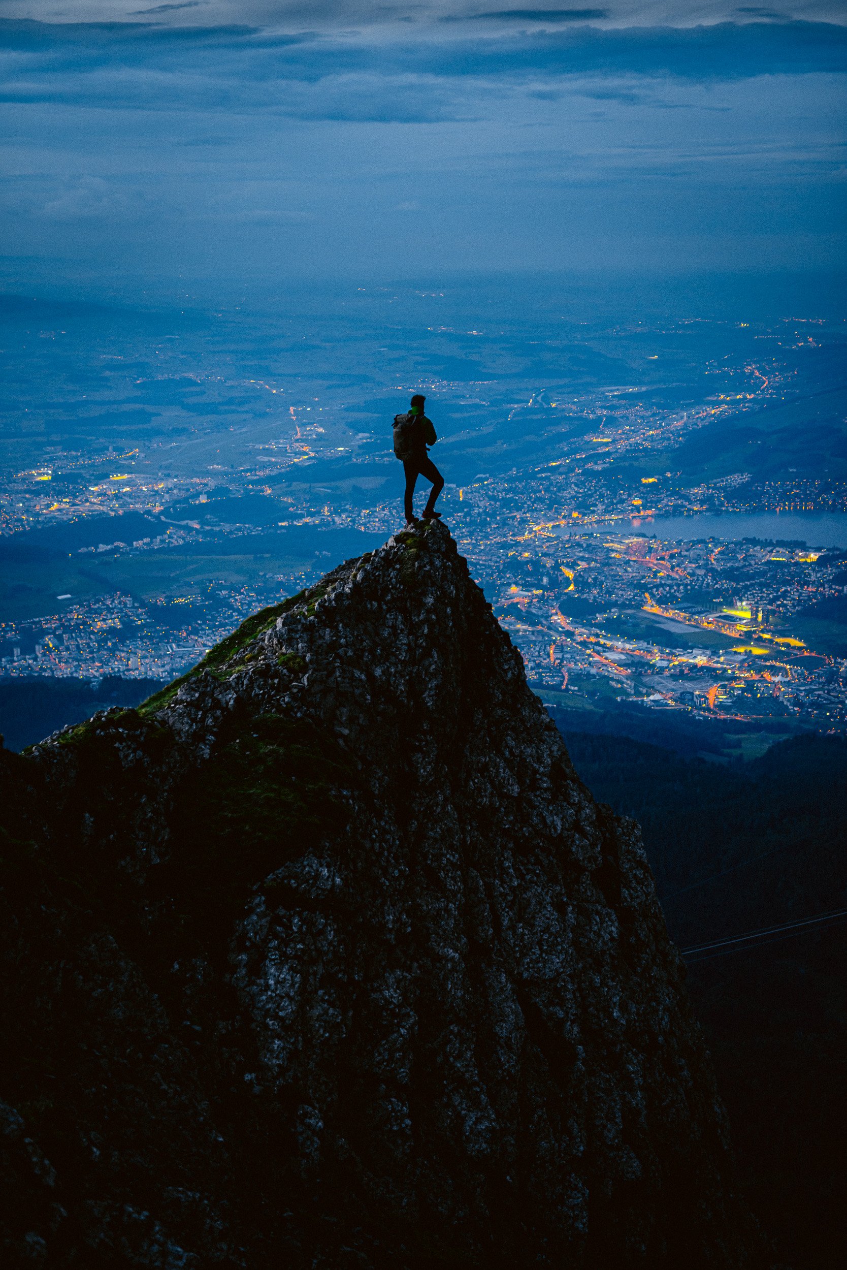 A person standing on a rocky mountain peak at dusk overlooking a valley with city lights.