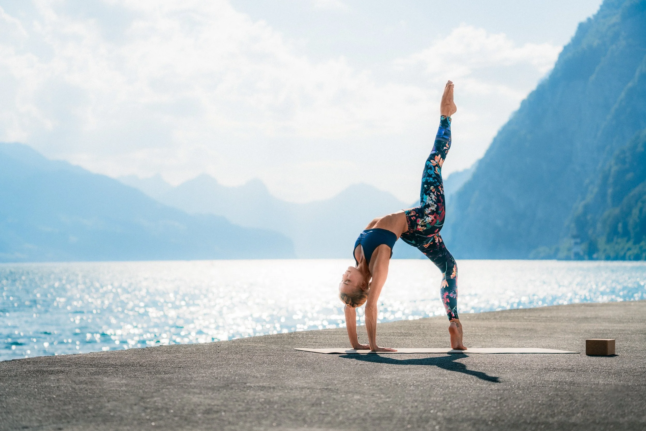 A woman practicing yoga outdoors on a mat by water, performing a one-legged downward dog pose with mountains in the background.