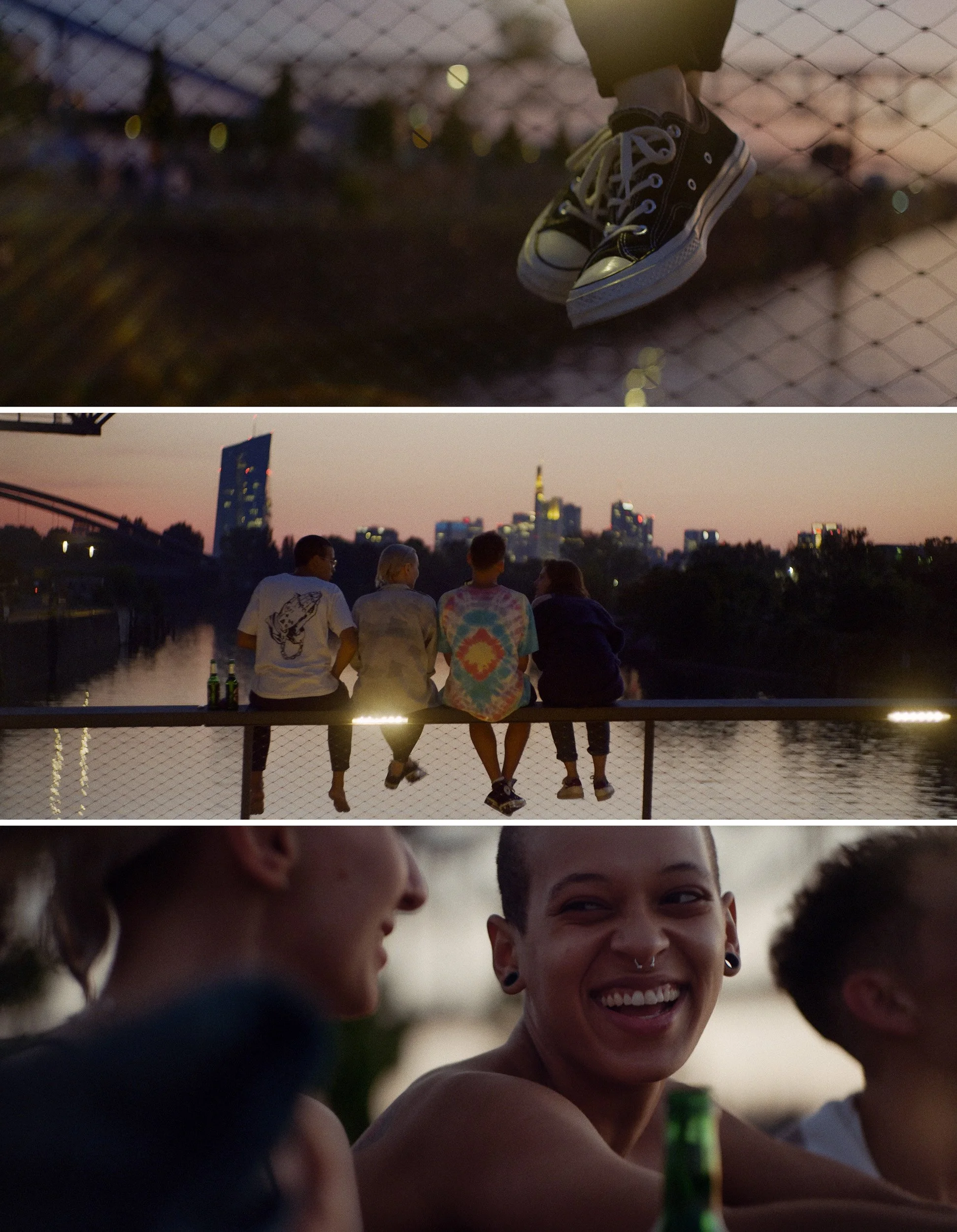 Three friends sitting on a railing by a river at sunset, chatting and smiling, with city skyline in the background.