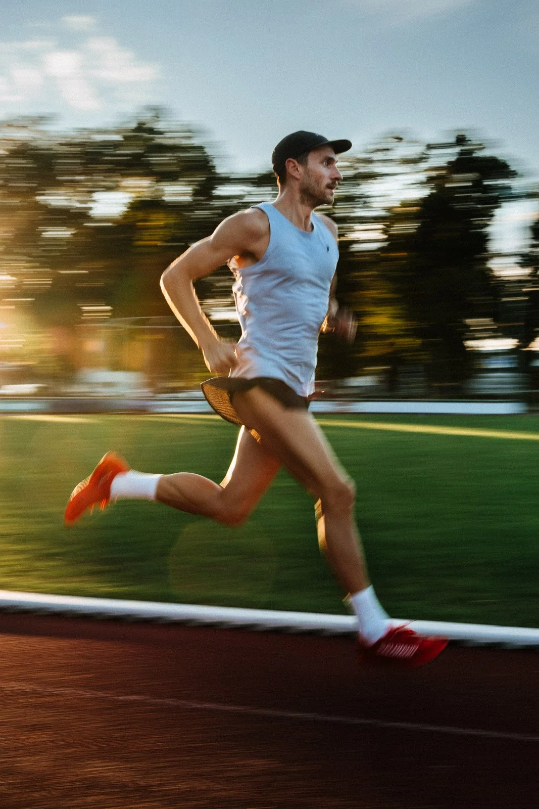 A man running outdoors on a track at sunset, wearing a gray tank top, black shorts, orange running shoes, and a black cap.
