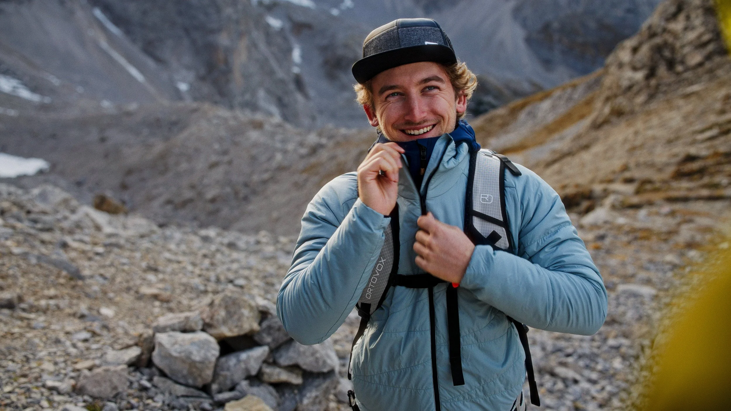 A smiling young man wearing a backpack, jacket, and cap, standing on rocky terrain in a mountainous landscape.