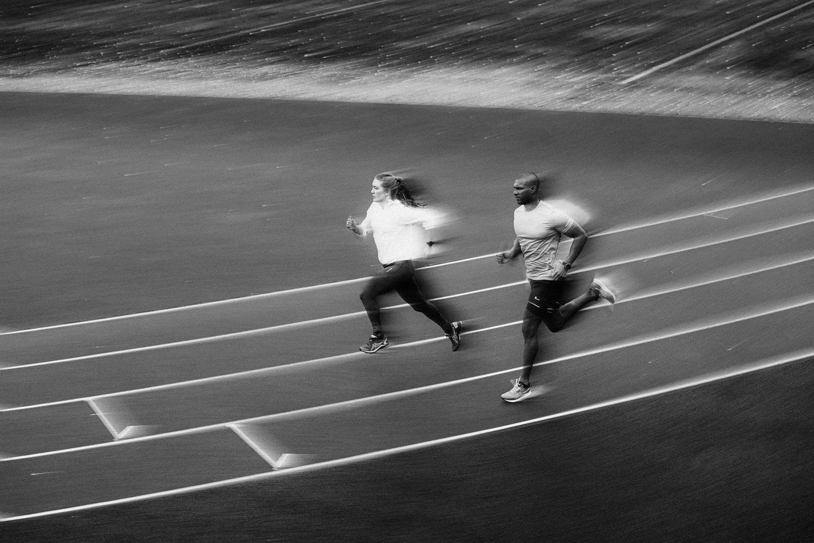 Black and white photo of two people running on a track in a stadium, with motion blur showing movement.