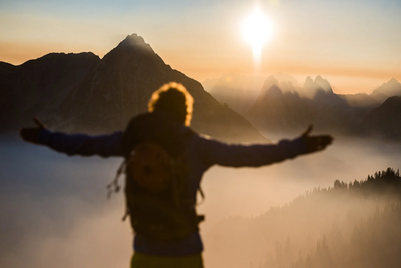 A person with a backpack standing with arms outstretched facing a mountain landscape during sunset.