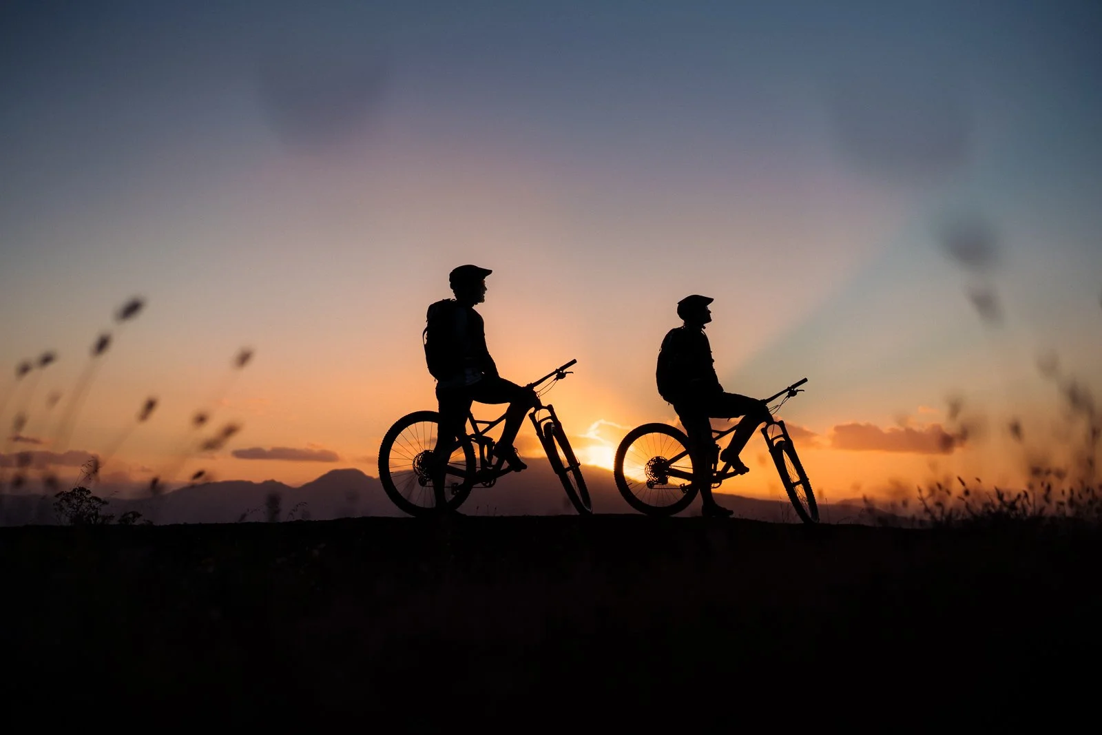Silhouettes of two people riding bicycles during sunset with a mountain range in the background.