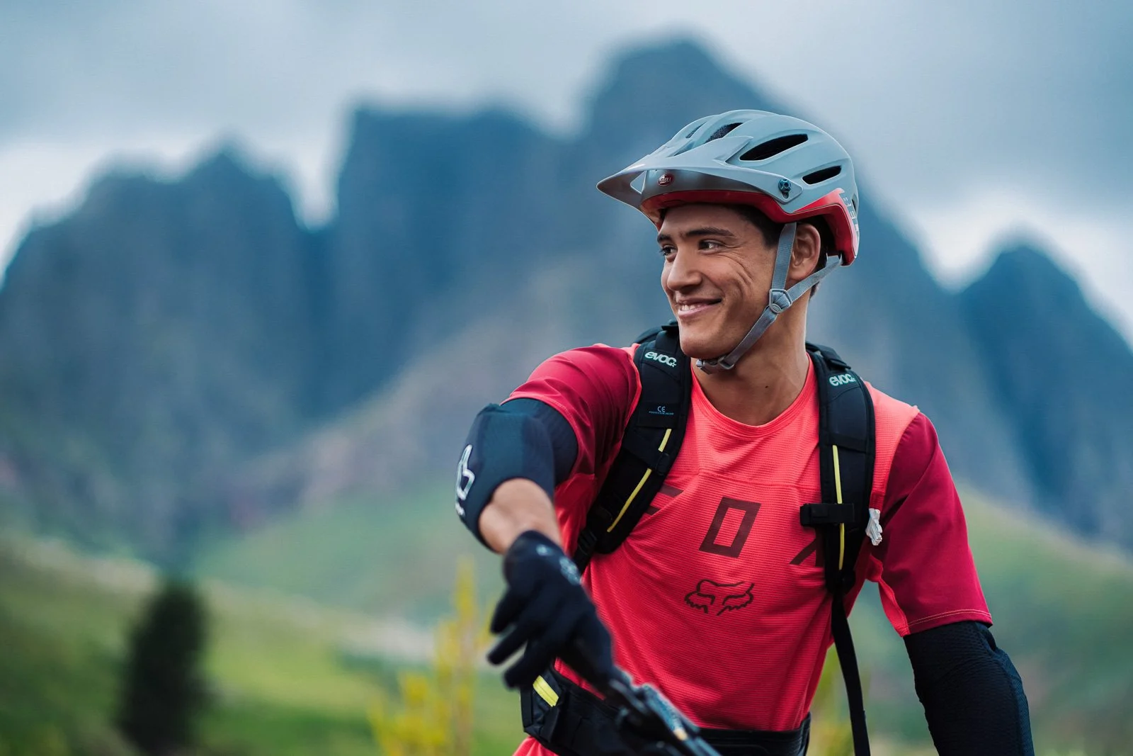 A man wearing a helmet and biking gear smiling outdoors with mountains in the background.