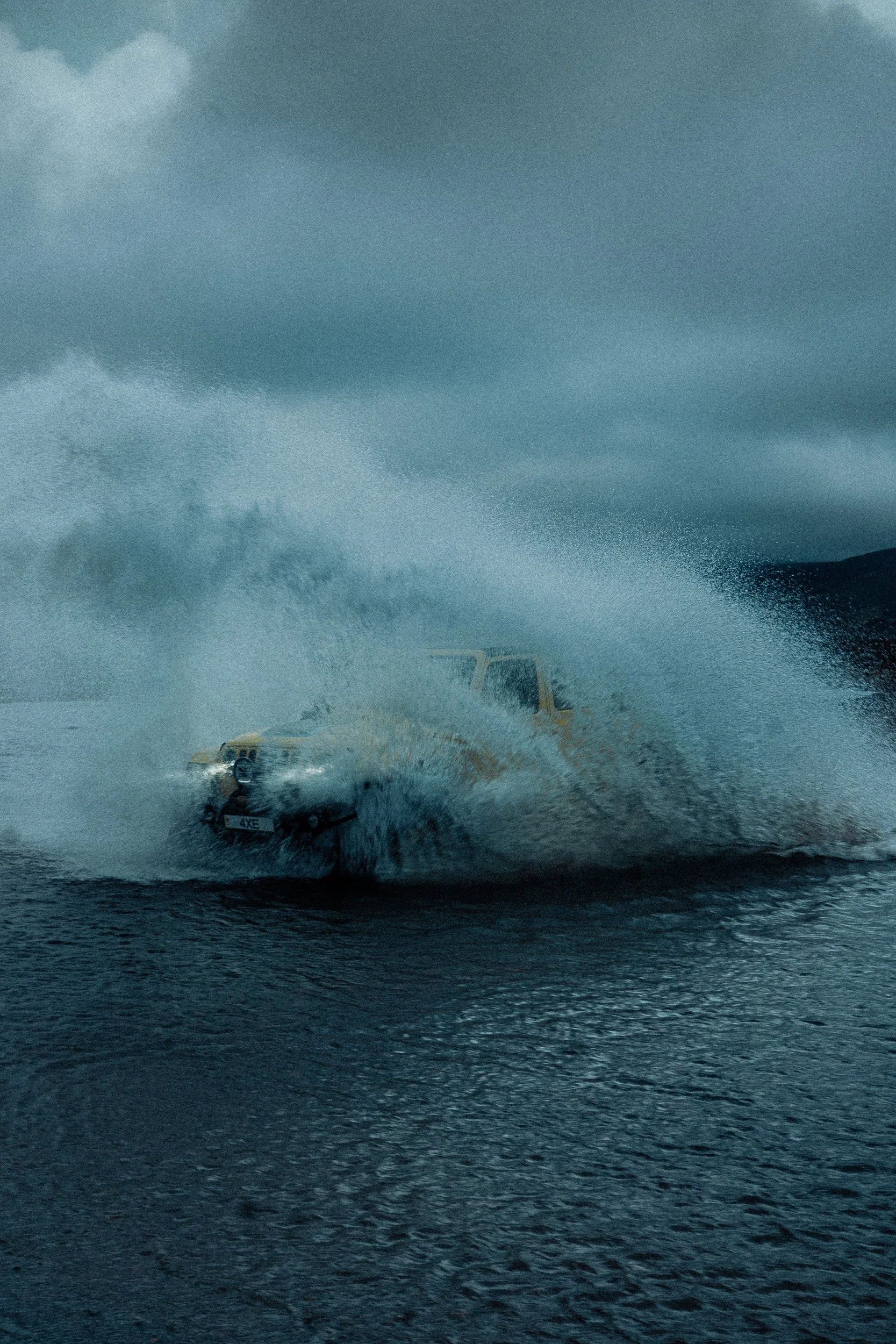 A yellow off-road vehicle driving through water, creating a large splash, with grey clouds in the sky.