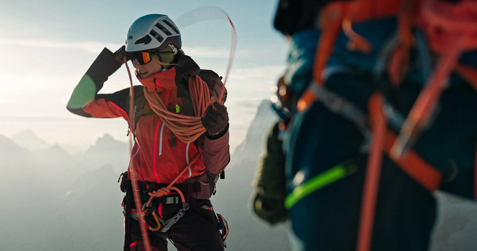 Two mountaineers in safety gear preparing to climb a snowy mountain, with one person adjusting their helmet and the other holding climbing equipment.