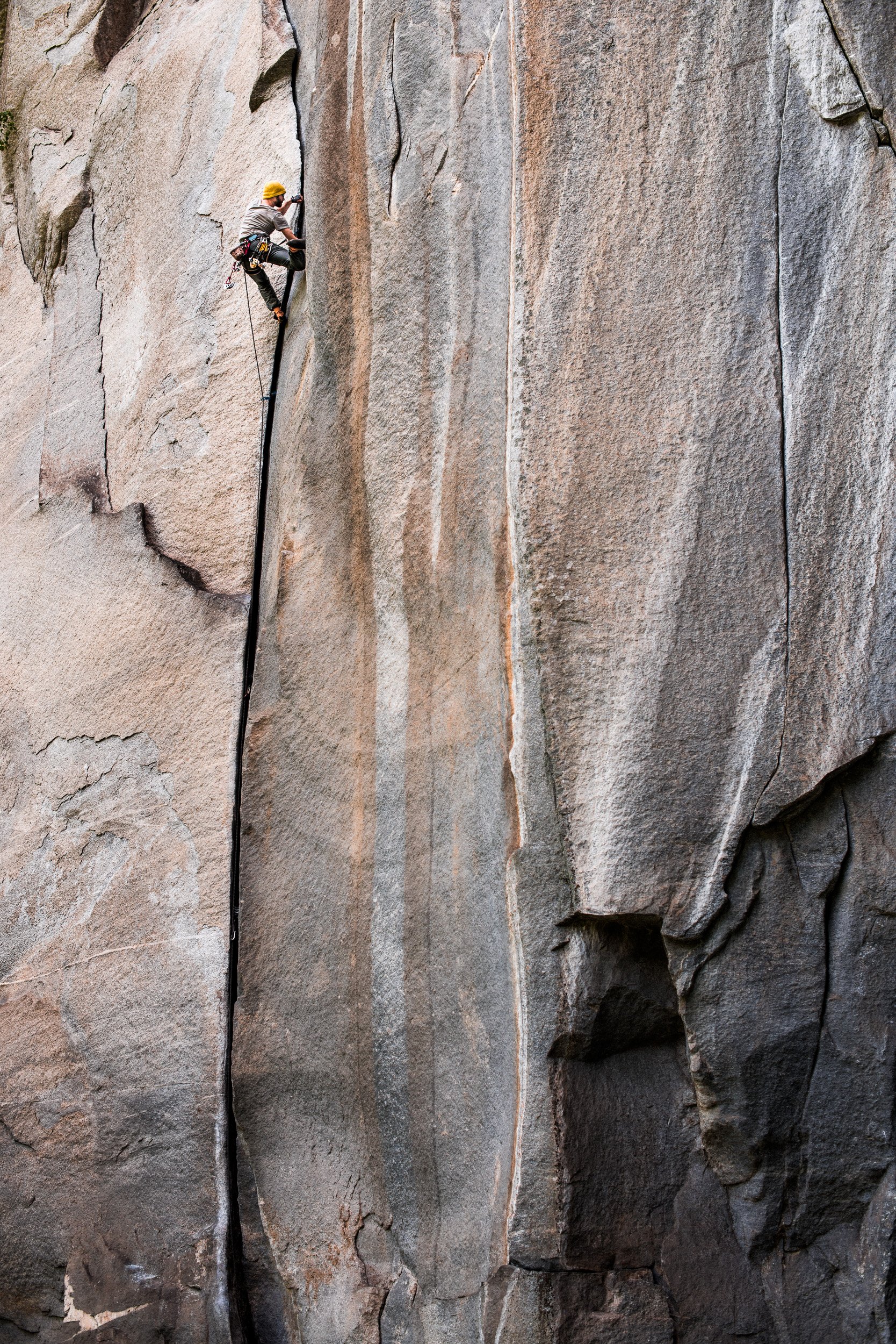 A person climbing a tall granite rock face with safety gear and a yellow helmet.