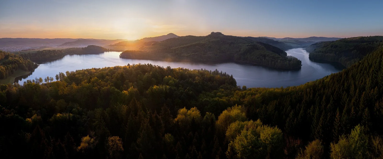 A scenic view of a winding river surrounded by dense forest under a sunset sky with hills and mountains in the distance.