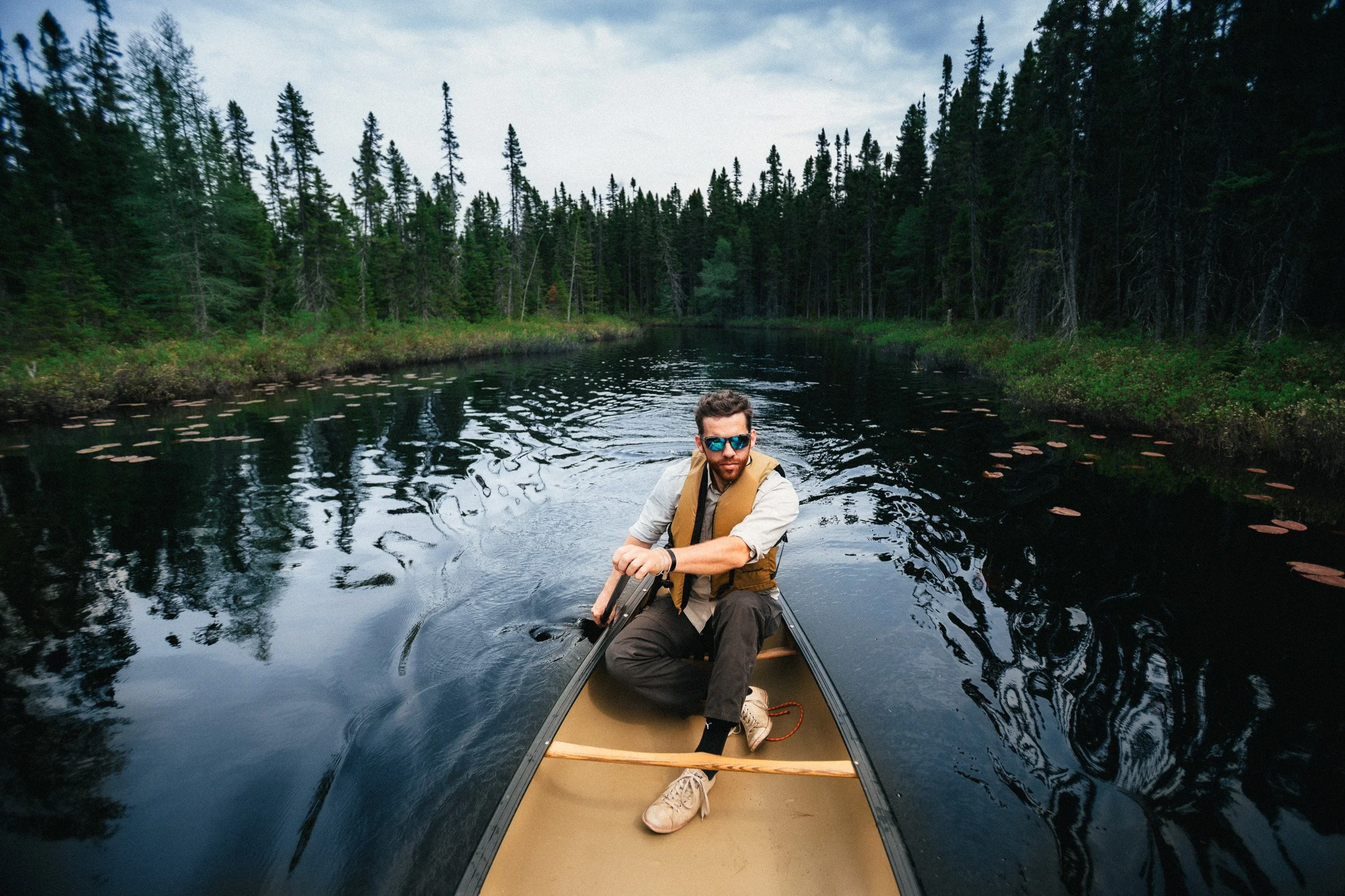 A man wearing sunglasses and a life jacket paddles a canoe on a calm river surrounded by dense green forests.