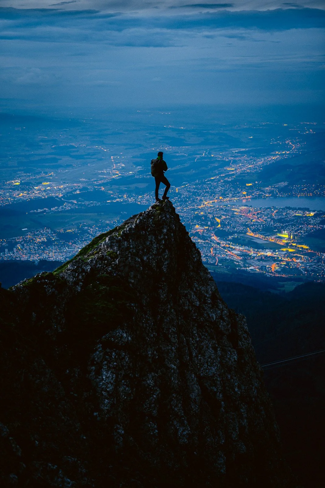 A person standing on the peak of a mountain during dusk, overlooking a city illuminated with lights below.