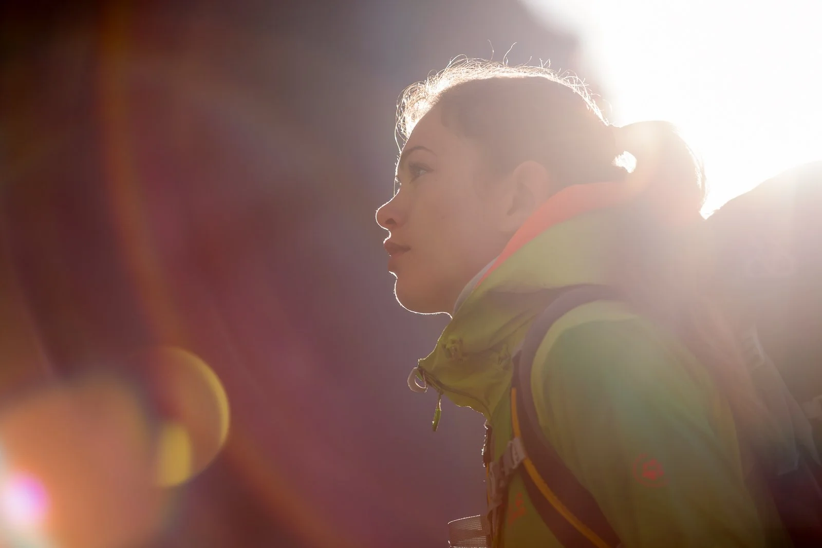 A woman with a ponytail outdoors in bright sunlight, wearing a yellow jacket and looking to the left.