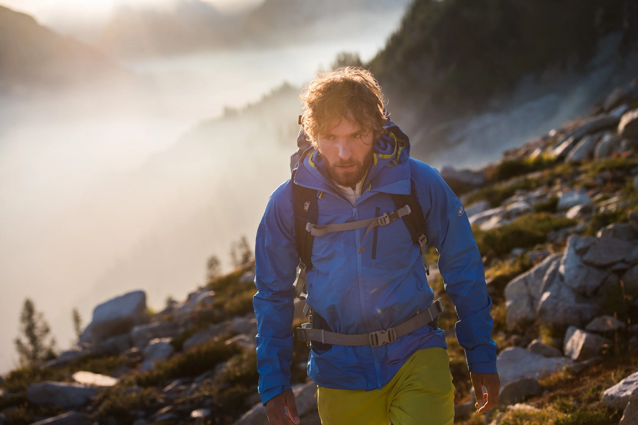 A man with curly hair and a beard hiking on a rocky mountain trail, wearing a blue jacket, yellow pants, and a backpack, with a background of misty mountains and a cloudy sky.