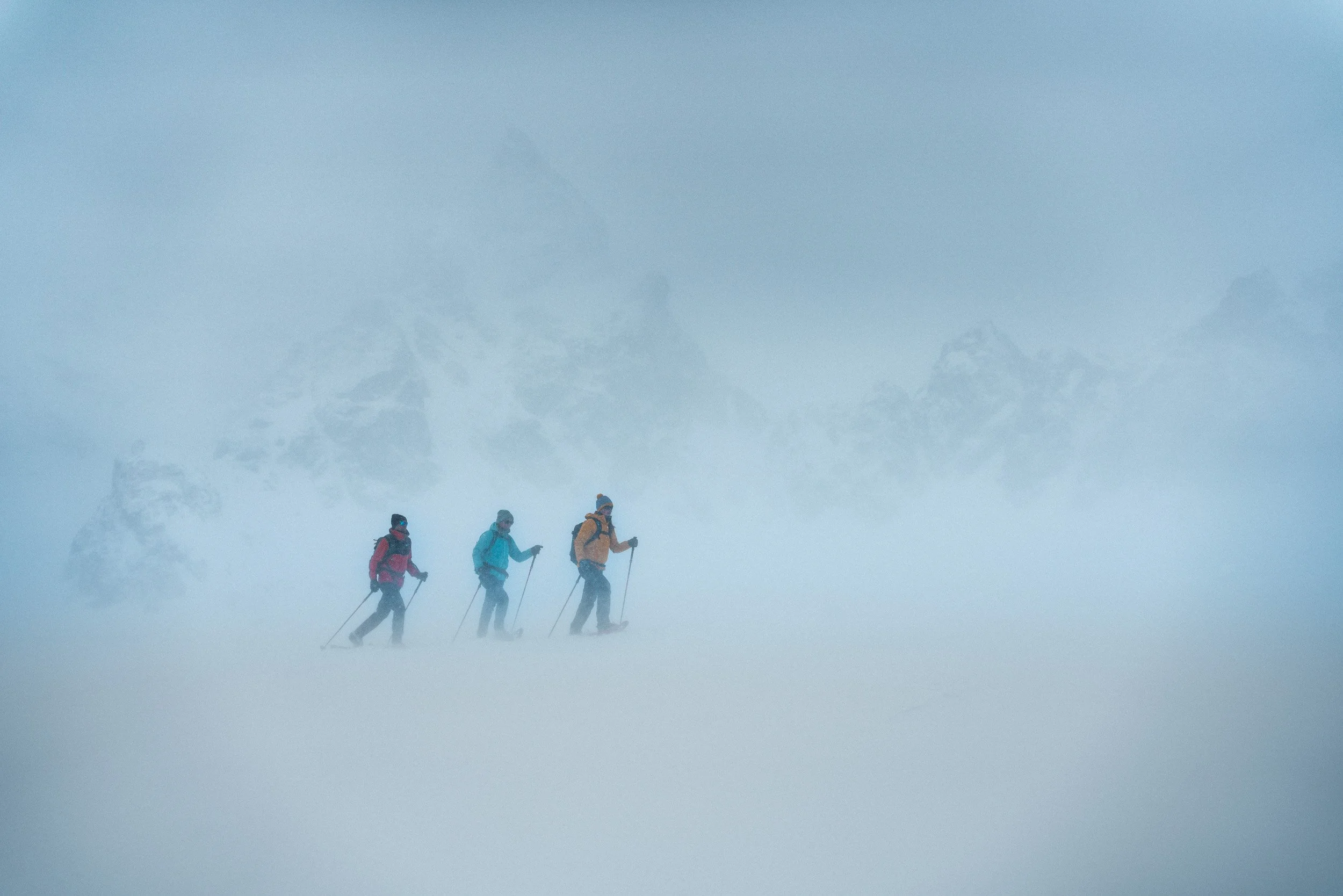Three hikers dressed in winter gear trekking through a snowy, foggy landscape with mountains in the background.