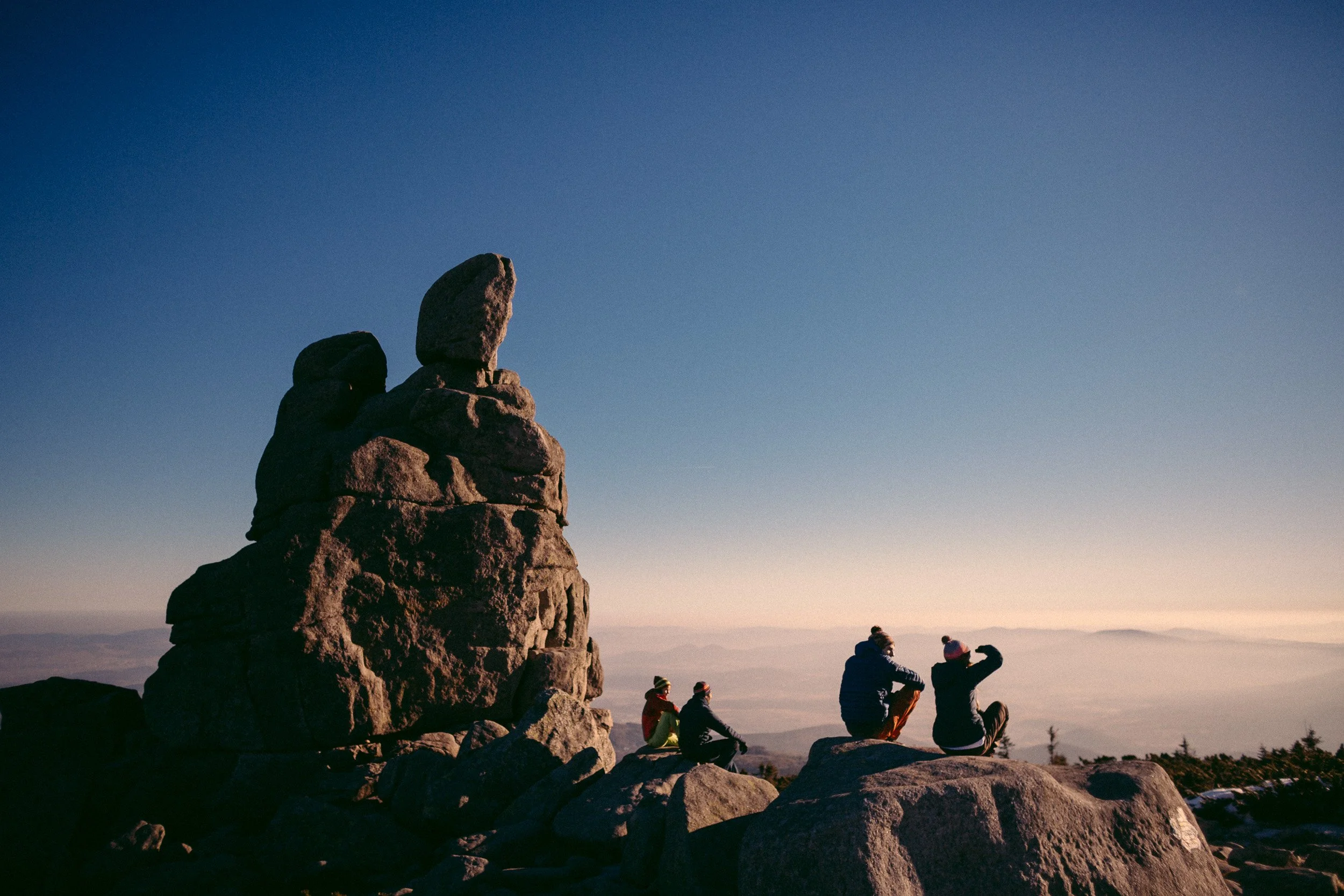 Group of four hikers sitting on rocks and watching the sunset over mountain landscape, with a large rock formation in the foreground.