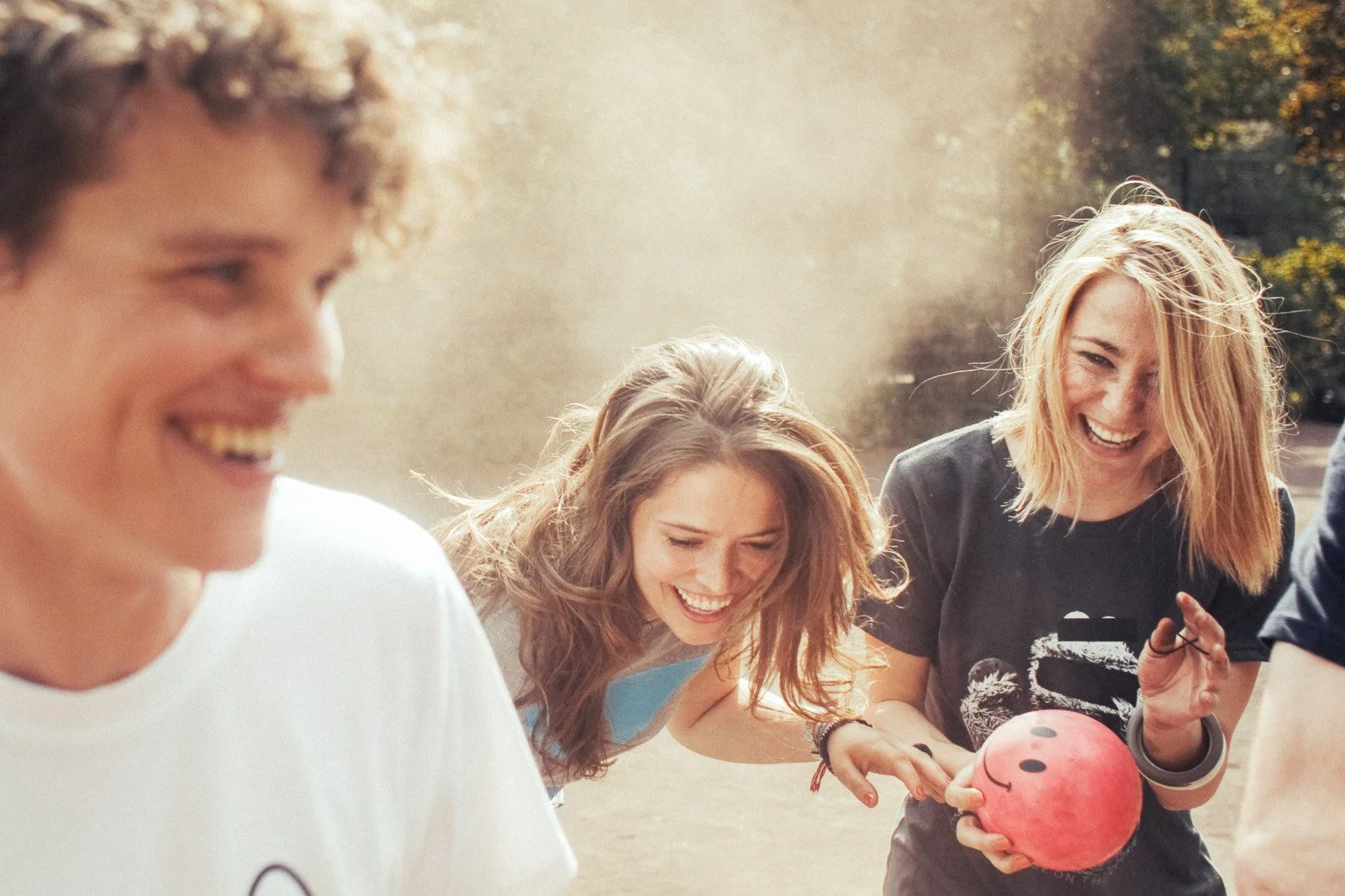 Three young women outdoors, laughing and playing with a pink bowling ball.